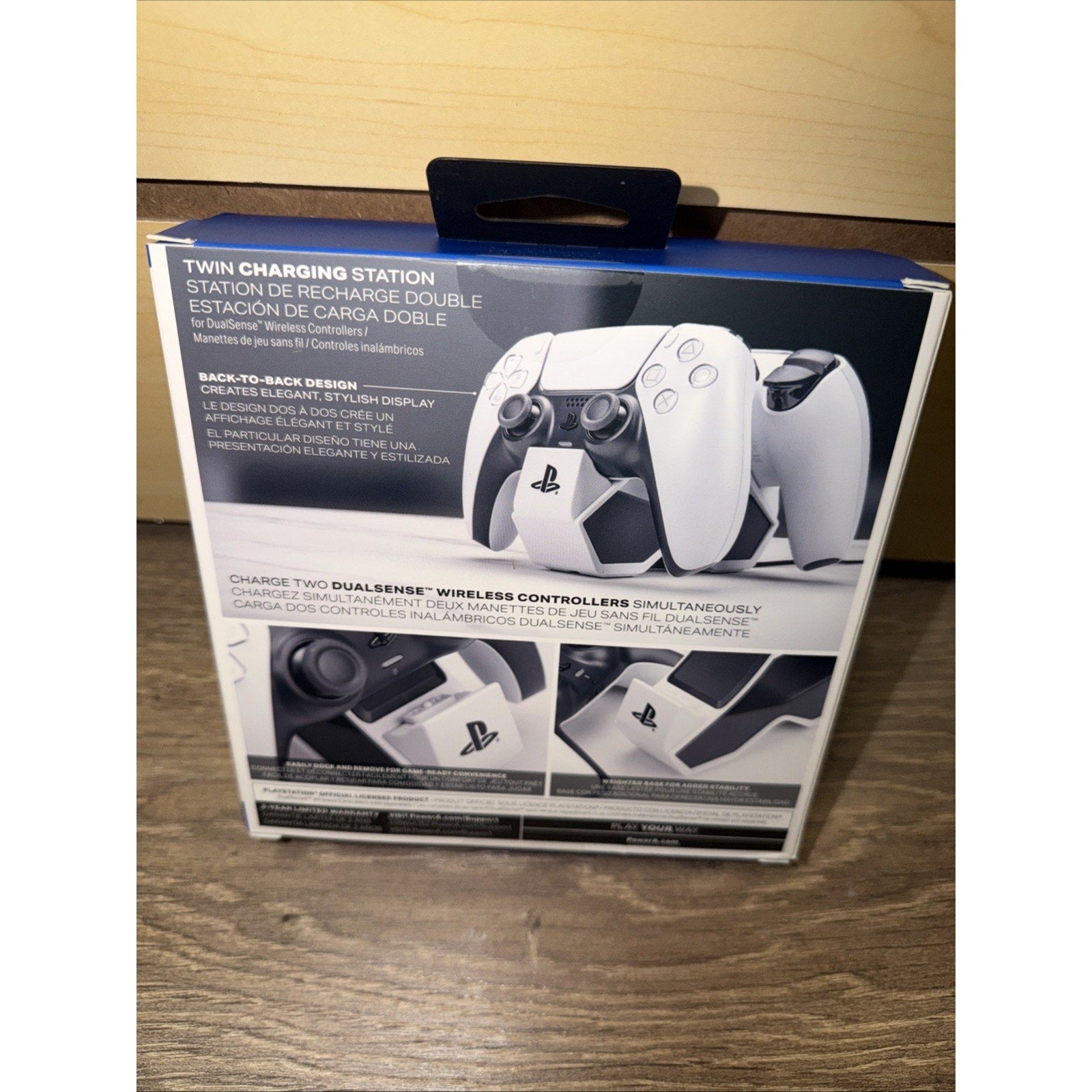 Packaging of a twin charging station for two white DualSense wireless controllers, featuring a back-to-back design and close-up views of the charging dock on a wooden surface.