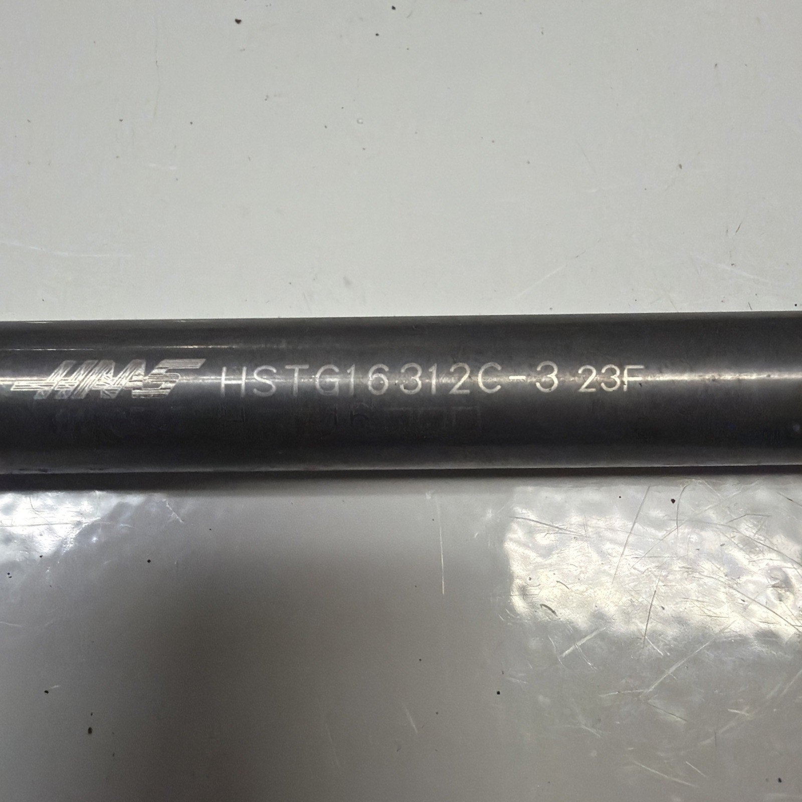 Black cylindrical metal rod with engraved alphanumeric code "HSTG16312C-3 23F" and faint brand text on a reflective white surface.