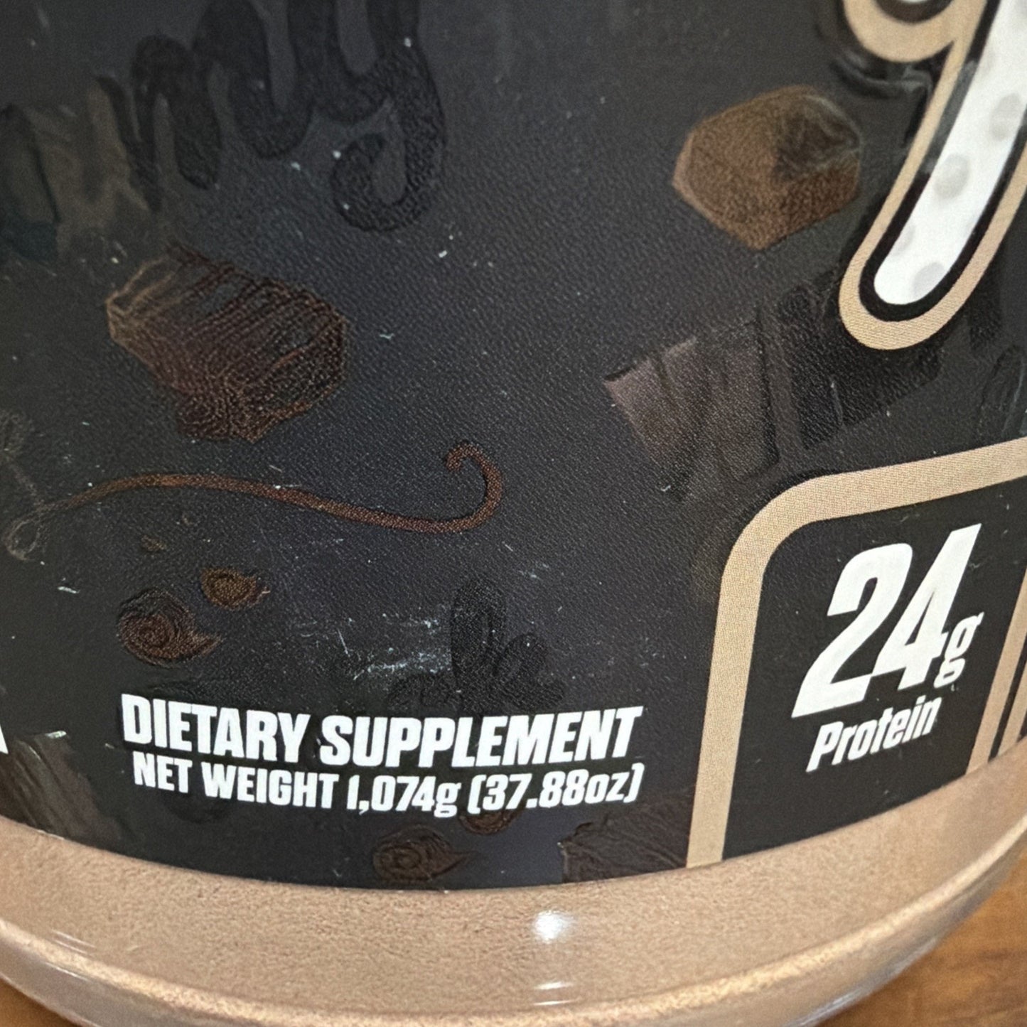 Close-up of a supplement container label showing "Dietary Supplement" text, net weight of 1,074 grams (37.88 ounces), and protein content of 24 grams with chocolate-themed background graphics.