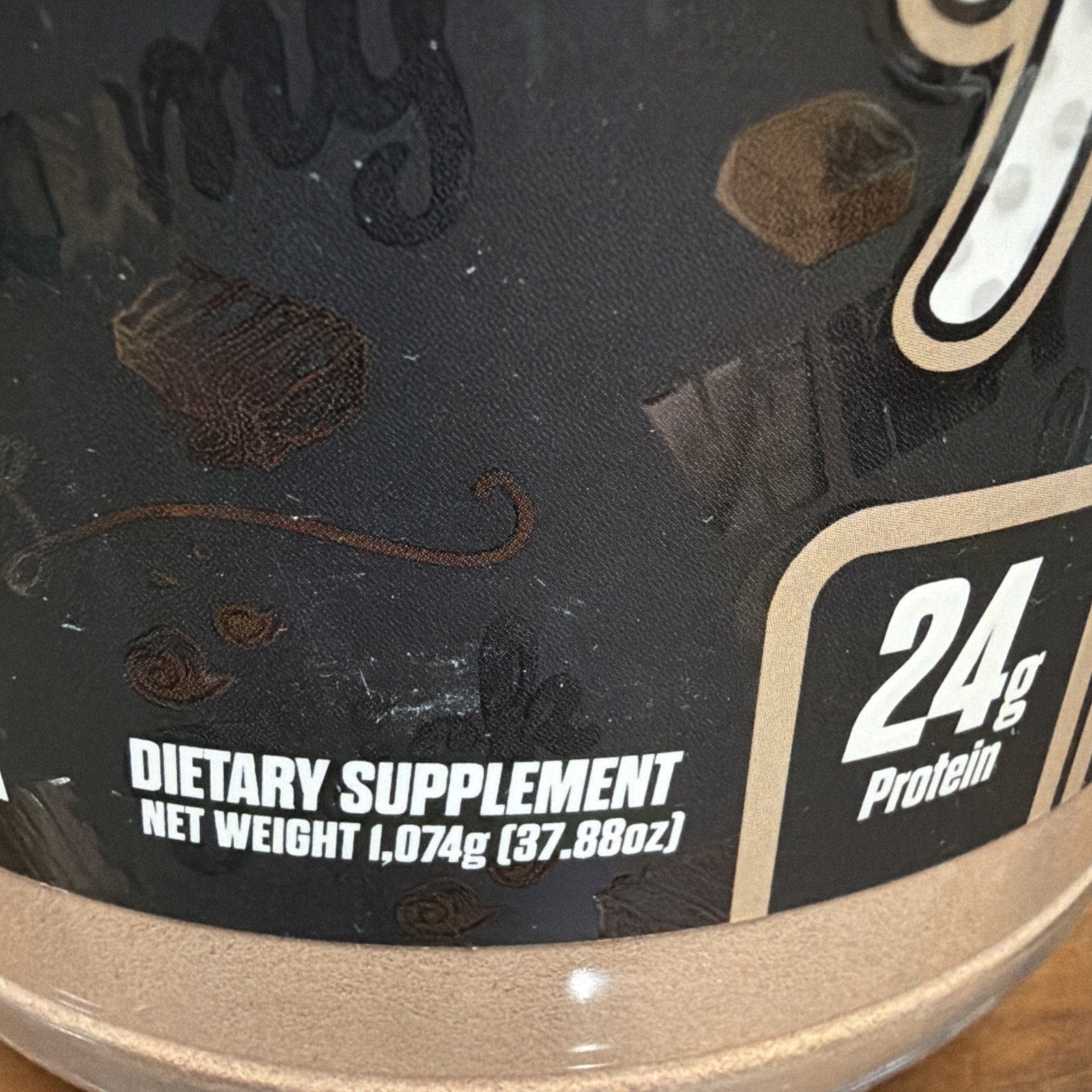 Close-up of a supplement container label showing "Dietary Supplement" text, net weight of 1,074 grams (37.88 ounces), and protein content of 24 grams with chocolate-themed background graphics.