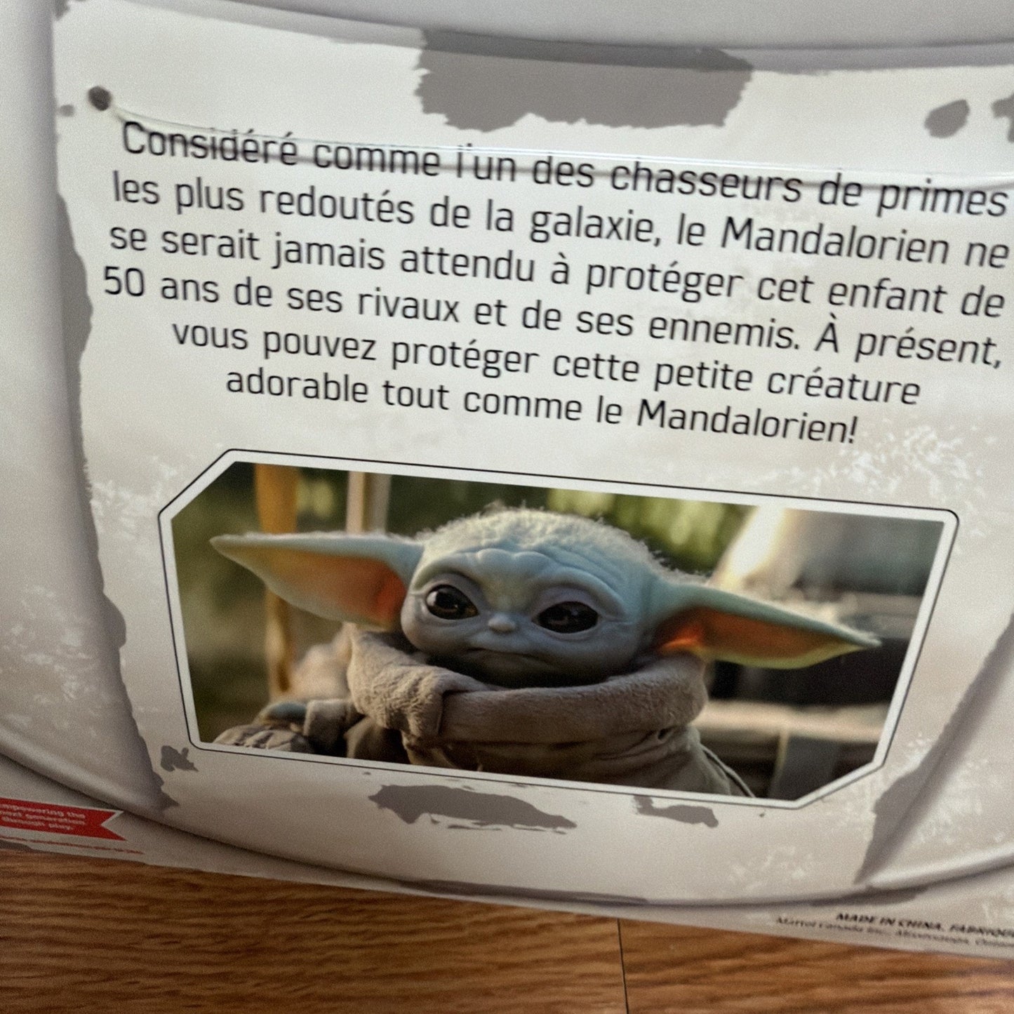 Close-up of a product label with French text describing the Mandalorian as a feared bounty hunter and encouraging protection of a small, adorable creature resembling Grogu from the Star Wars series, accompanied by an image of the character with large ears and big eyes wrapped in a brown robe.