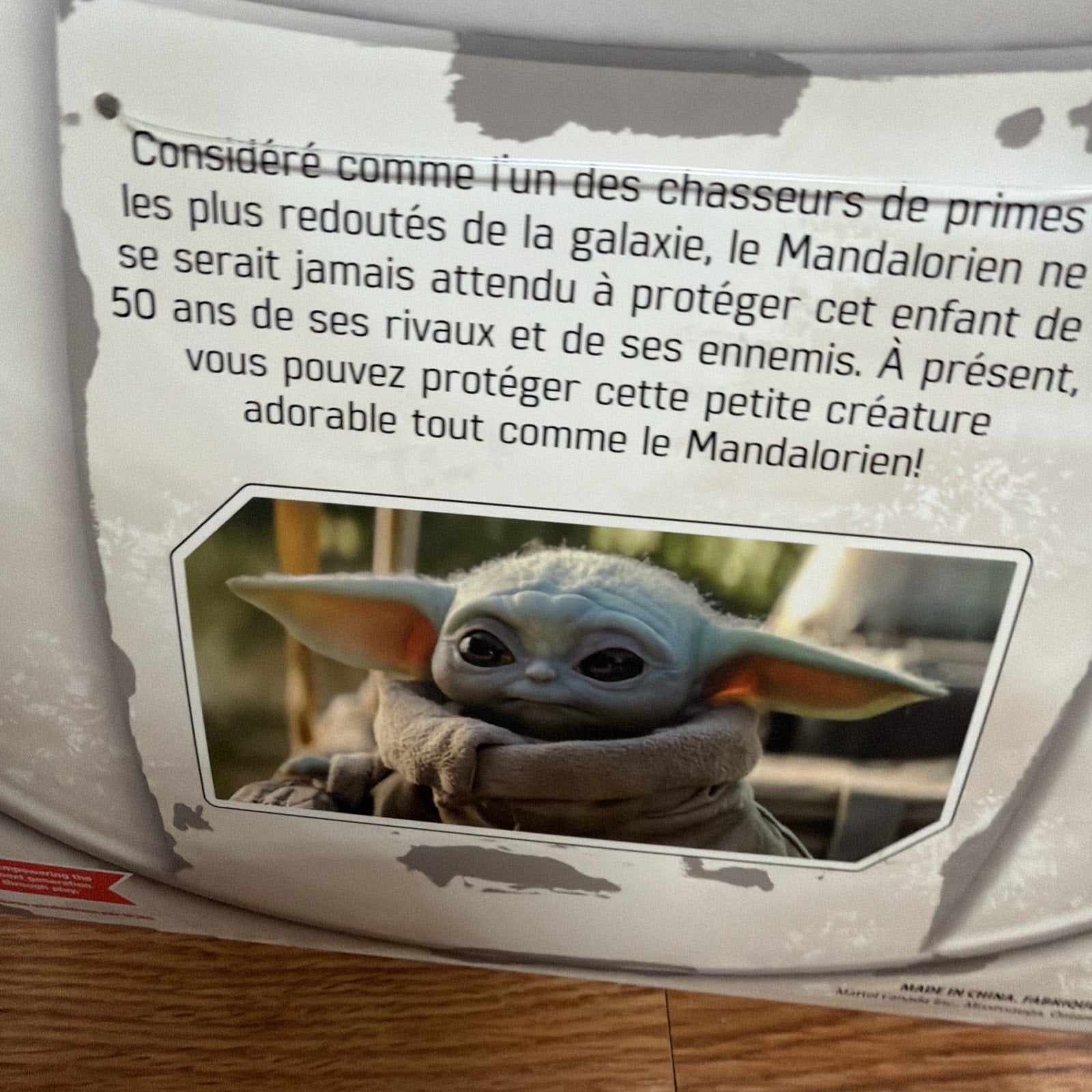 Close-up of a product label with French text describing the Mandalorian as a feared bounty hunter and encouraging protection of a small, adorable creature resembling Grogu from the Star Wars series, accompanied by an image of the character with large ears and big eyes wrapped in a brown robe.