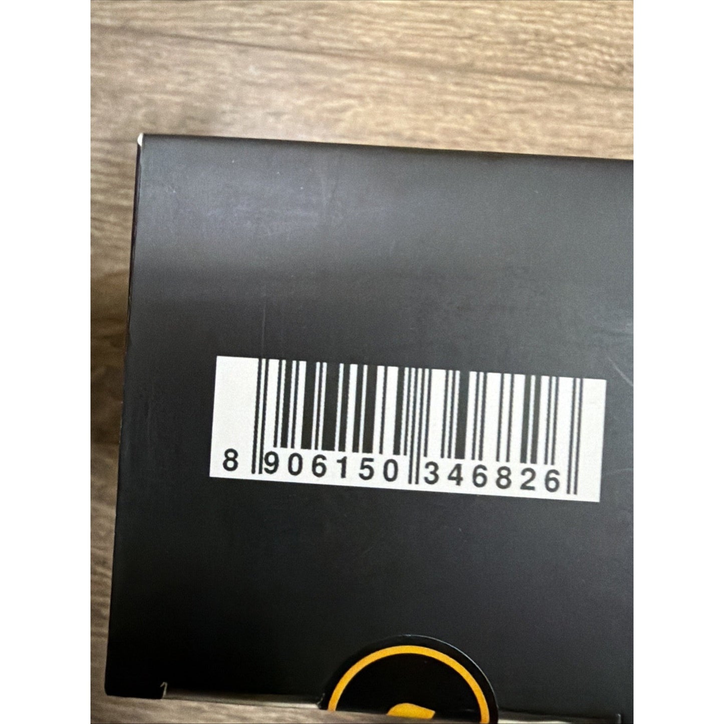 Close-up of a white barcode sticker with black lines and numbers on the matte black surface of a product box placed on a light wooden background.