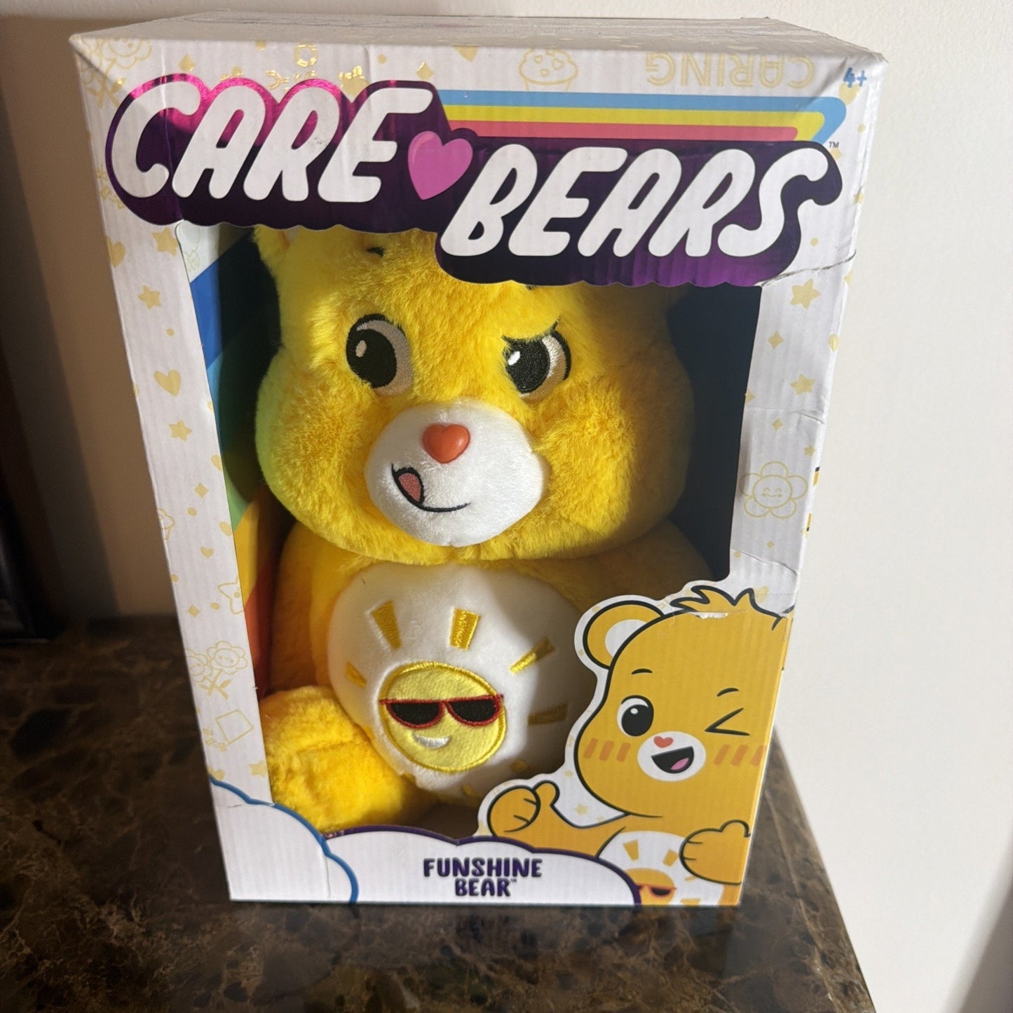 Yellow Care Bears plush toy with a sun wearing sunglasses emblem on its belly, packaged in a box featuring colorful Care Bears logo and cartoon character illustration.