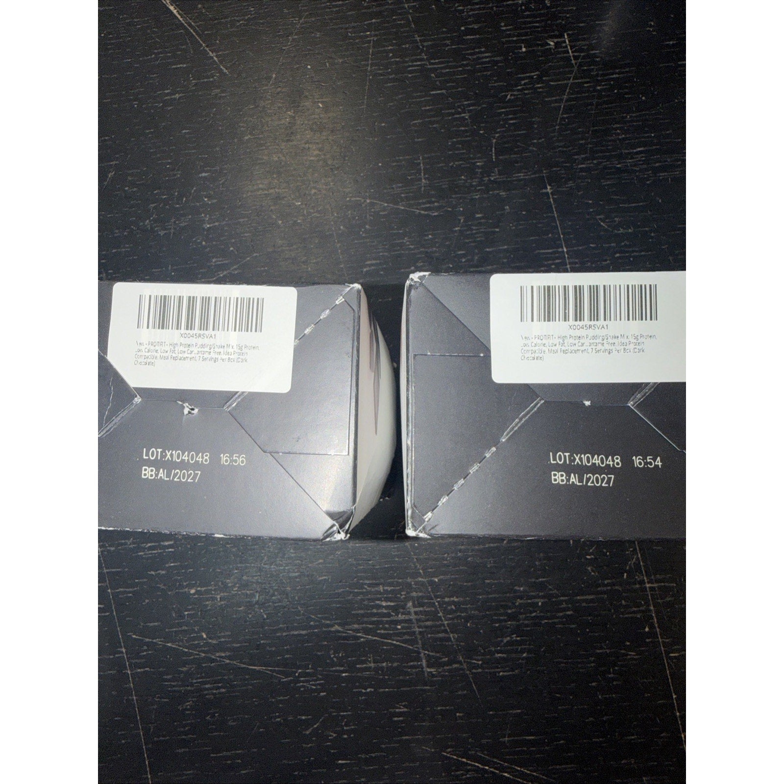 Two dark gray product boxes with white barcode labels and printed lot numbers placed on a scratched black surface.