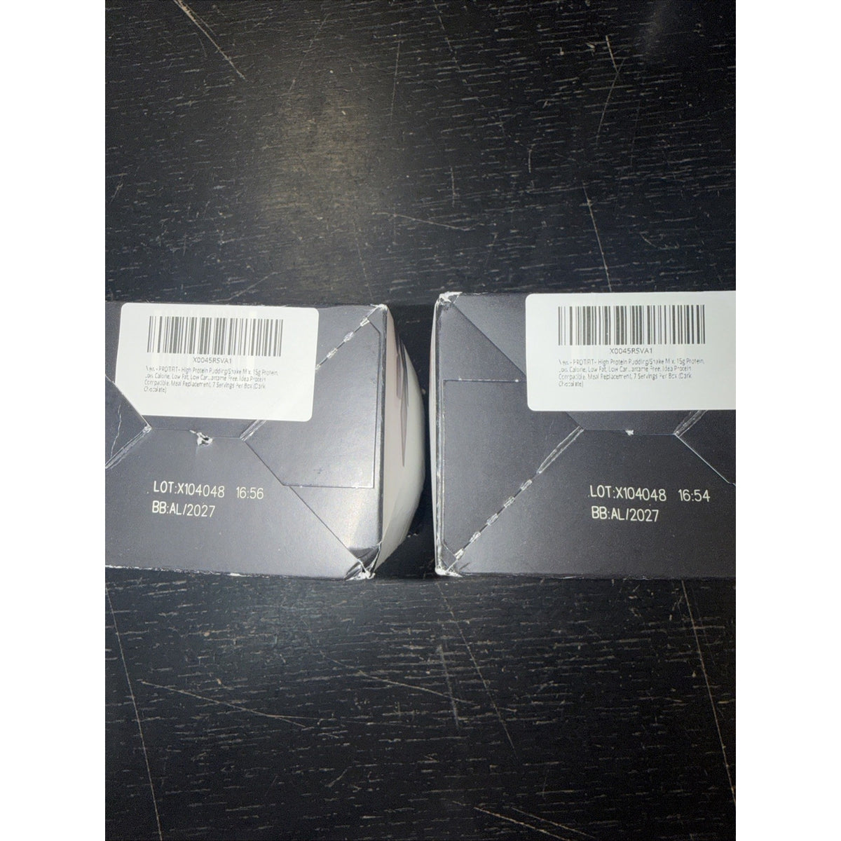 Two dark gray product boxes with white barcode labels and printed lot numbers placed on a scratched black surface.
