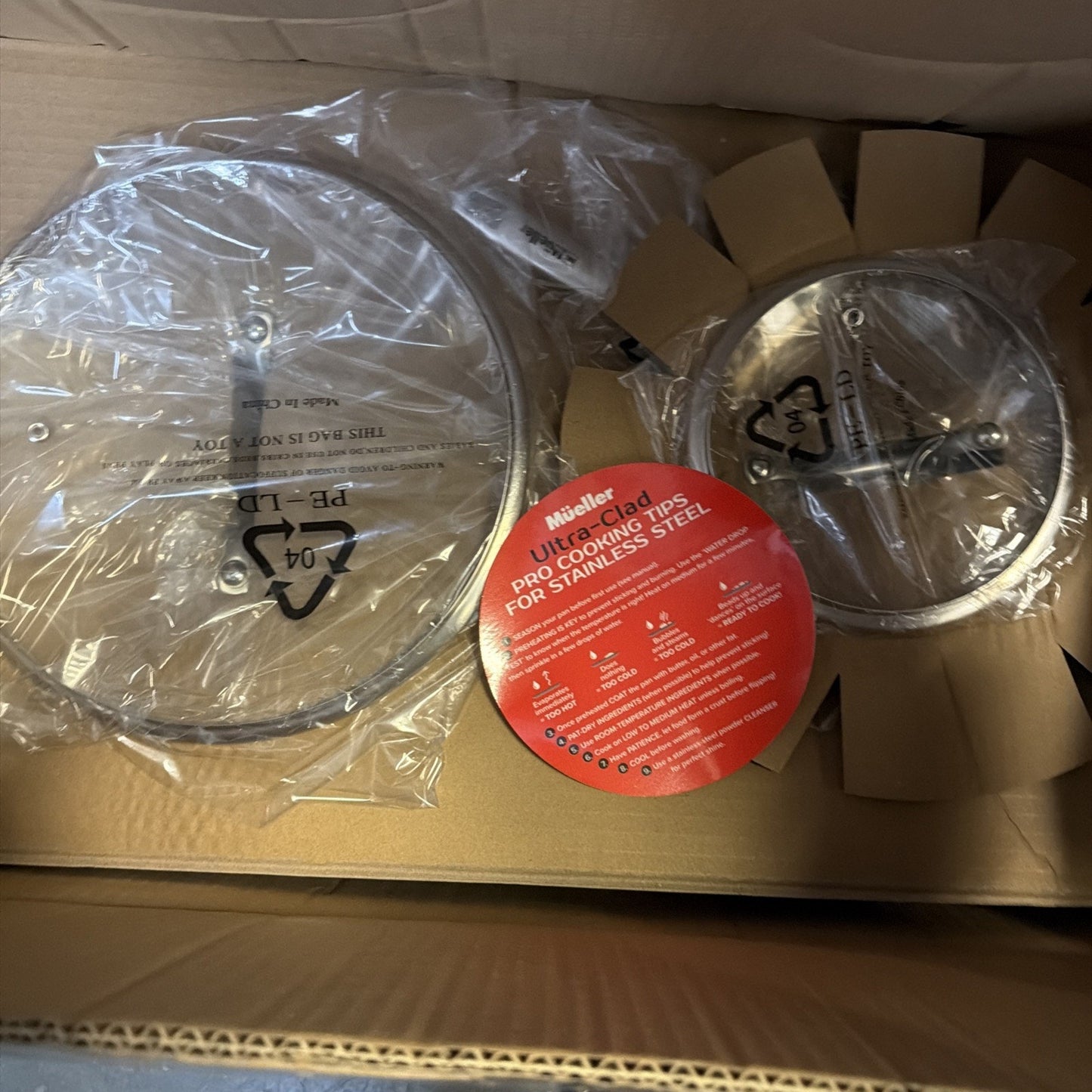 Two stainless steel cookware lids wrapped in plastic inside a cardboard box, one featuring a red label with cooking tips from Mueller Ultra-Clad.