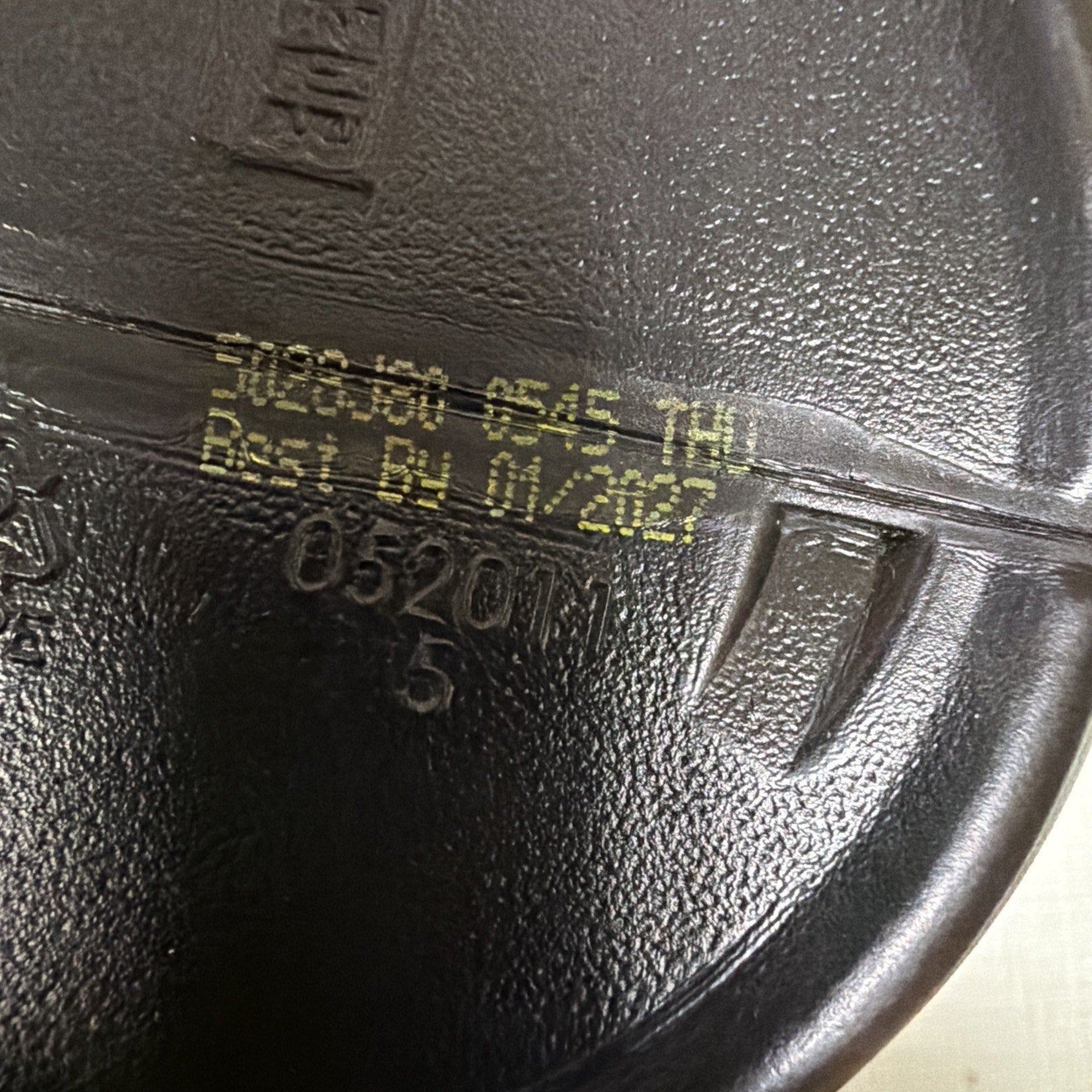 Close-up of a black plastic container bottom showing yellow printed text with batch code "318-133 6545 THU" and expiration date "Best By 01/2027" on a textured surface.