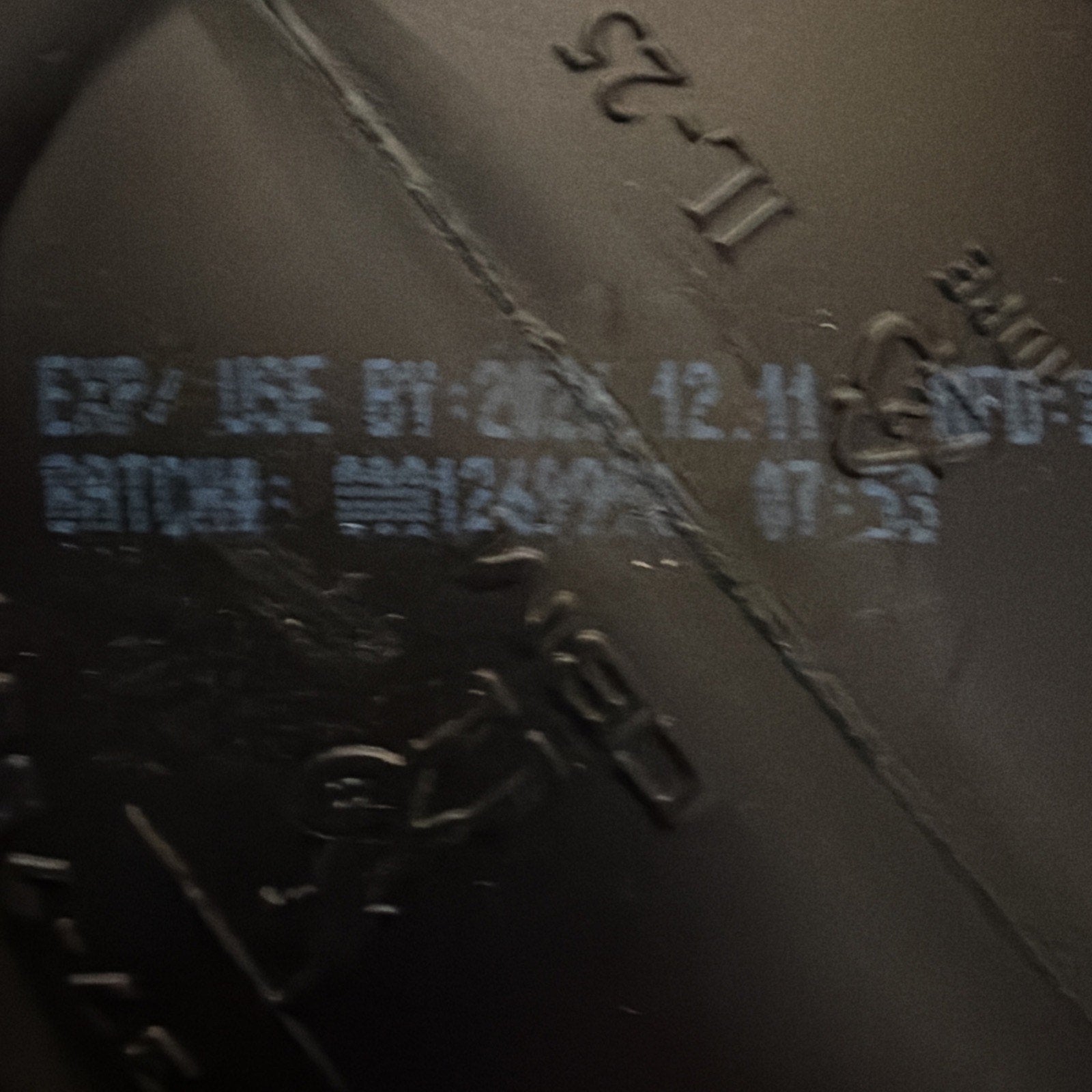 Close-up of a black plastic surface with light blue printed expiration date "2023.12.11" and batch code details, including time stamp "07:53" on a textured background.