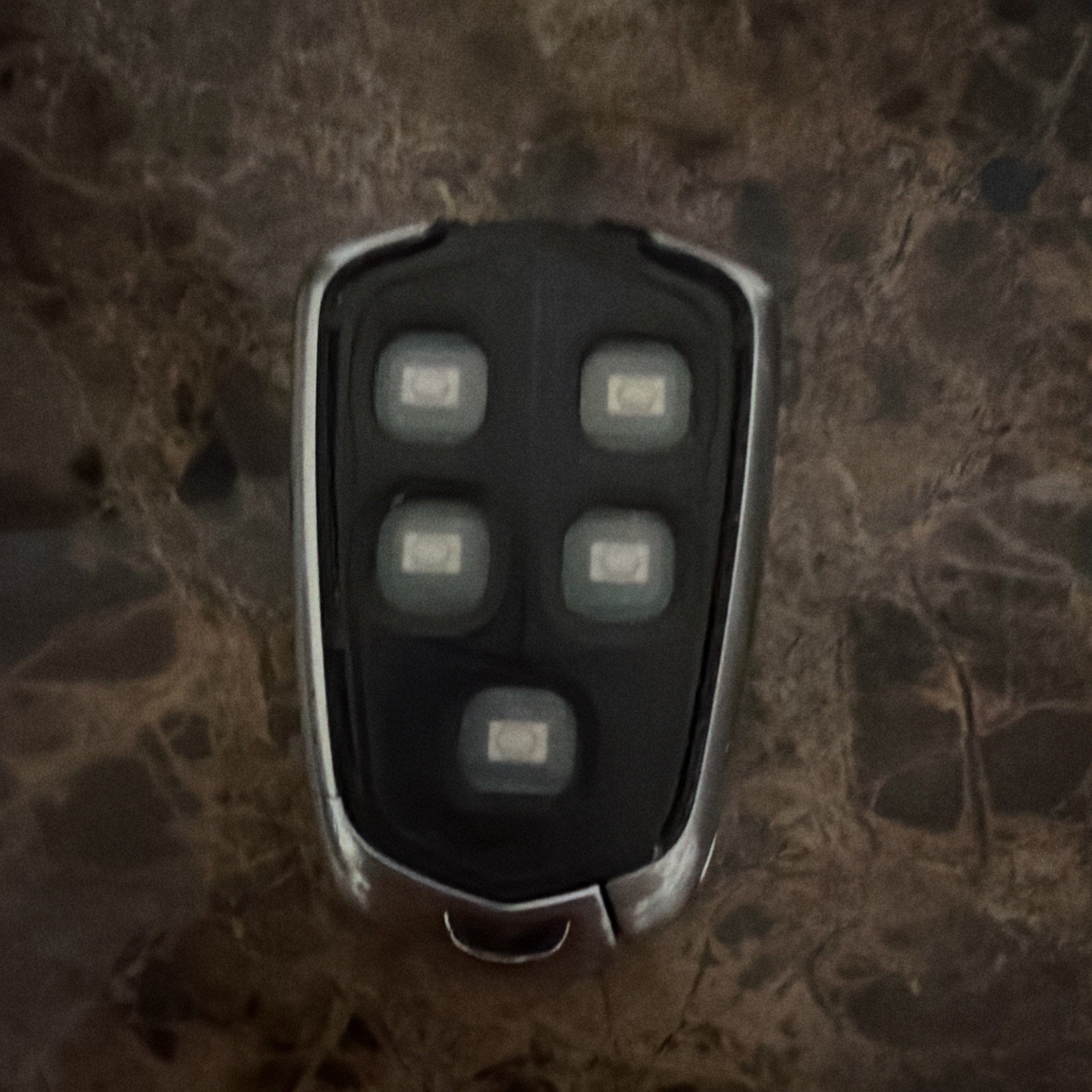 Close-up of a black car key fob with five rubberized buttons set in a chrome-trimmed casing, placed on a dark marbled surface.