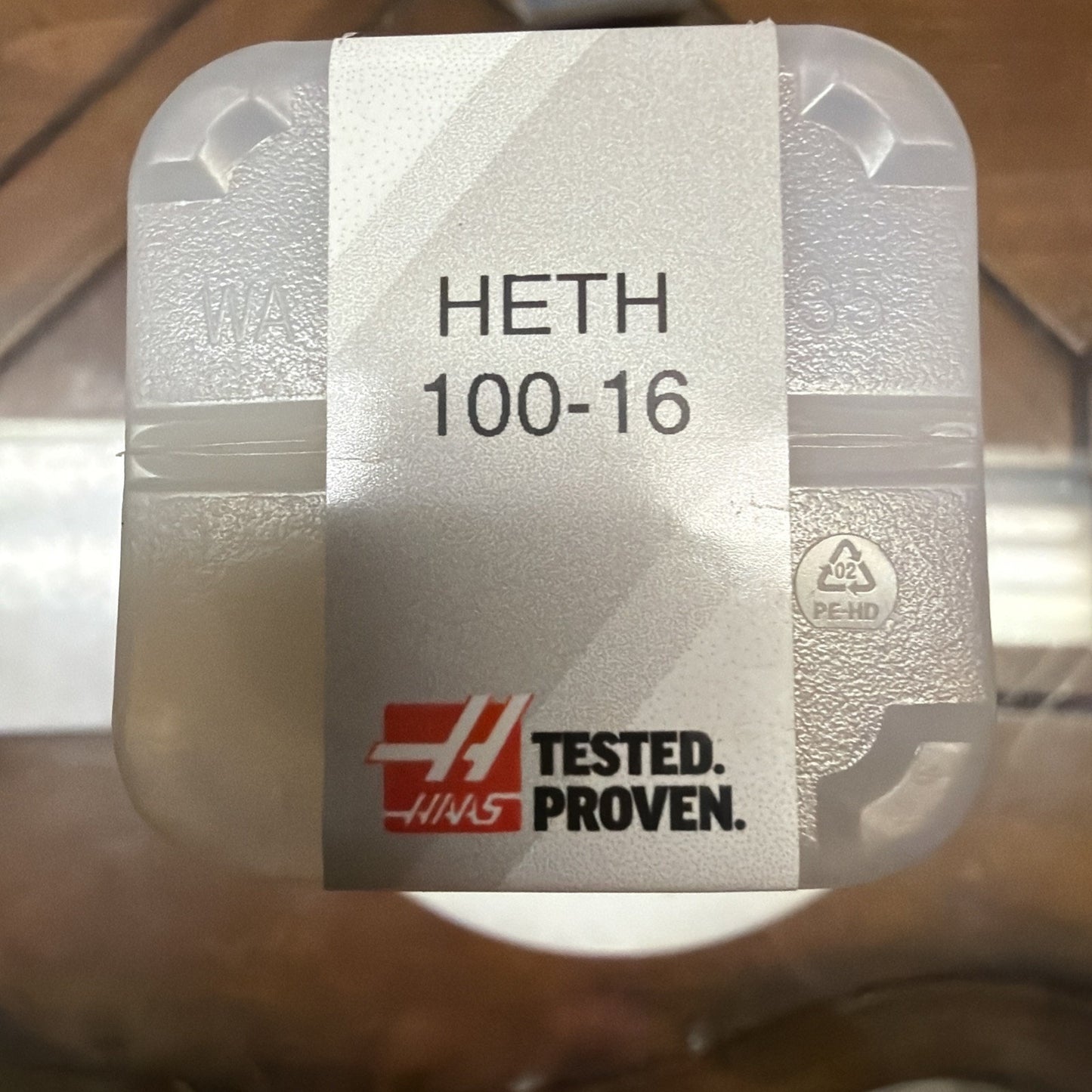 White textured plastic container with a label reading "HETH 100-16" and a red logo featuring the text "TESTED. PROVEN." on a wooden surface background.