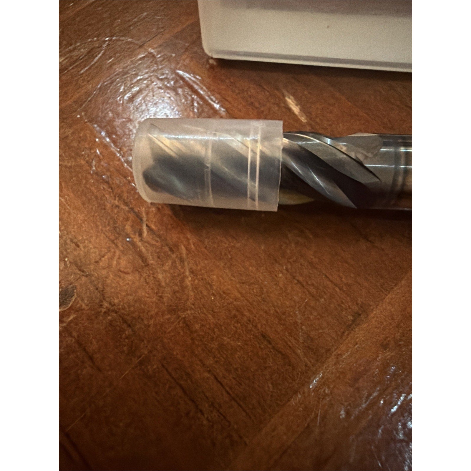 Close-up of a metal drill bit with spiral flutes partially covered by a translucent plastic protective cap, resting on a wooden surface with visible grain and scratches.