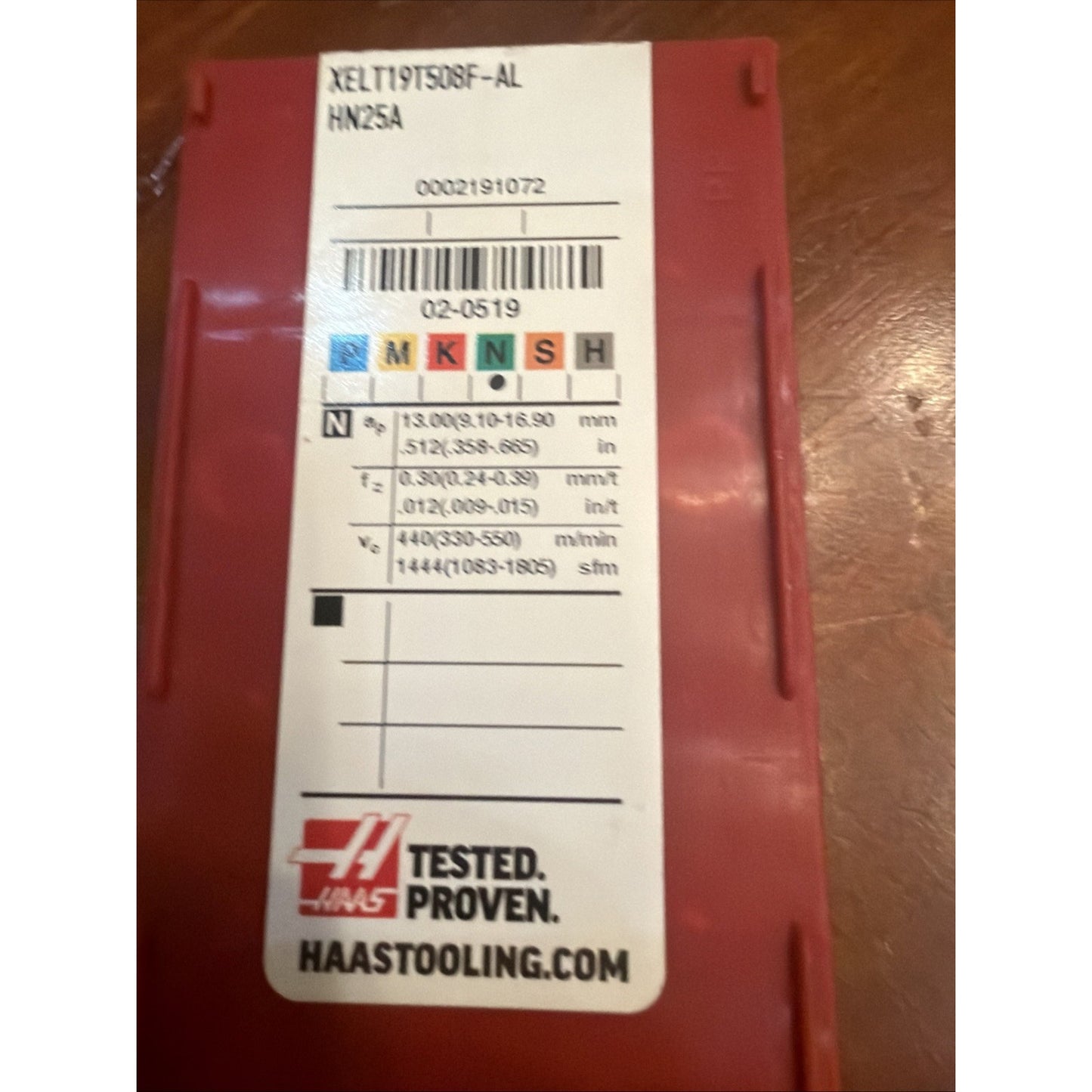Red plastic storage case with a white label displaying product code "XELTY9T50SF-AL HN25A," machining parameters, colored ISO classification squares, barcode, Haas logo, and URL, placed on a wooden surface.
