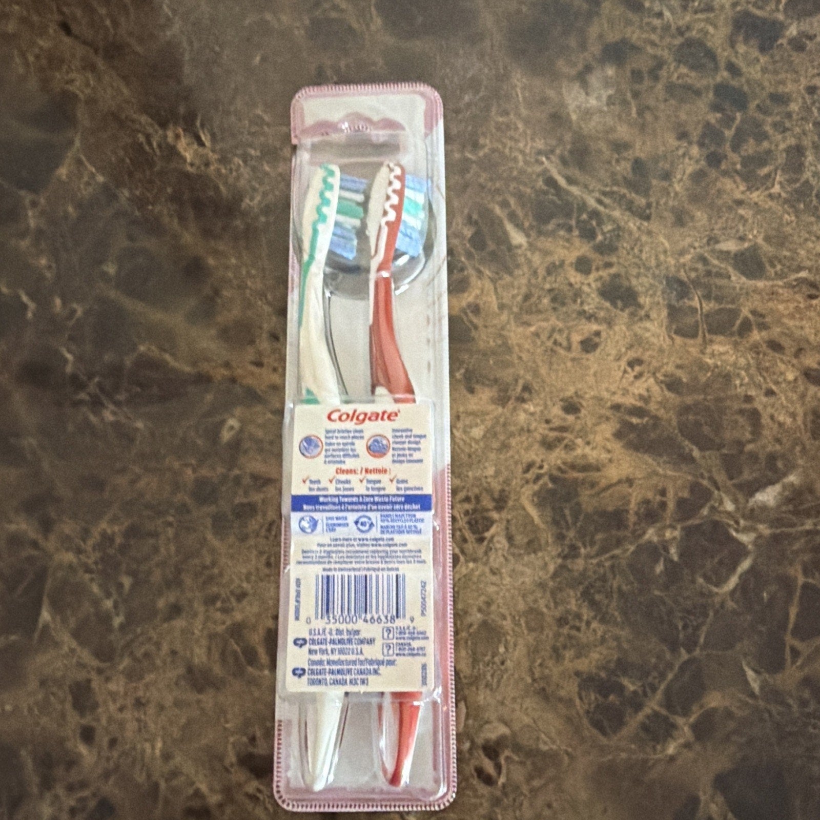 Two Colgate toothbrushes in clear plastic packaging with blue, white, and red handles visible on a brown marble surface, showing the back label and barcode.