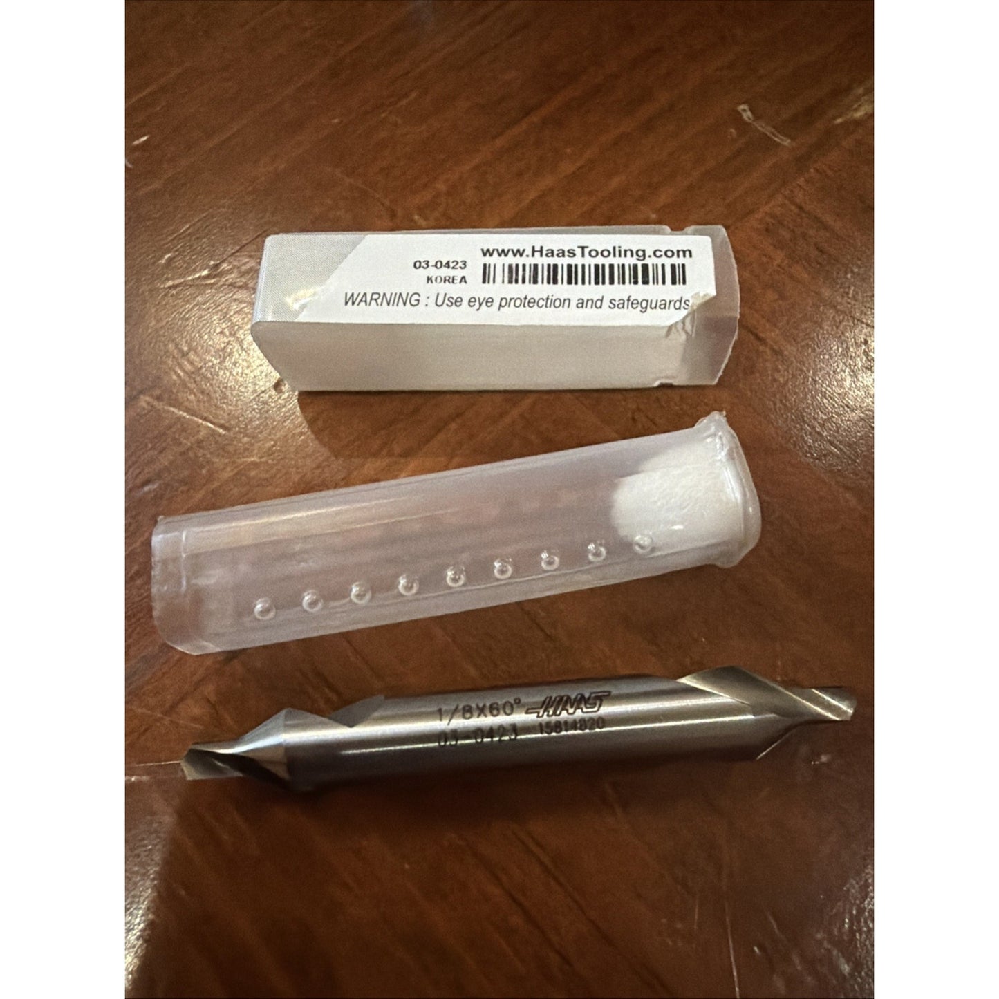 Metal drill bit with double-ended cutting edges, marked with size information, placed on a wooden surface alongside a clear plastic protective case and a white rectangular box with a warning label.