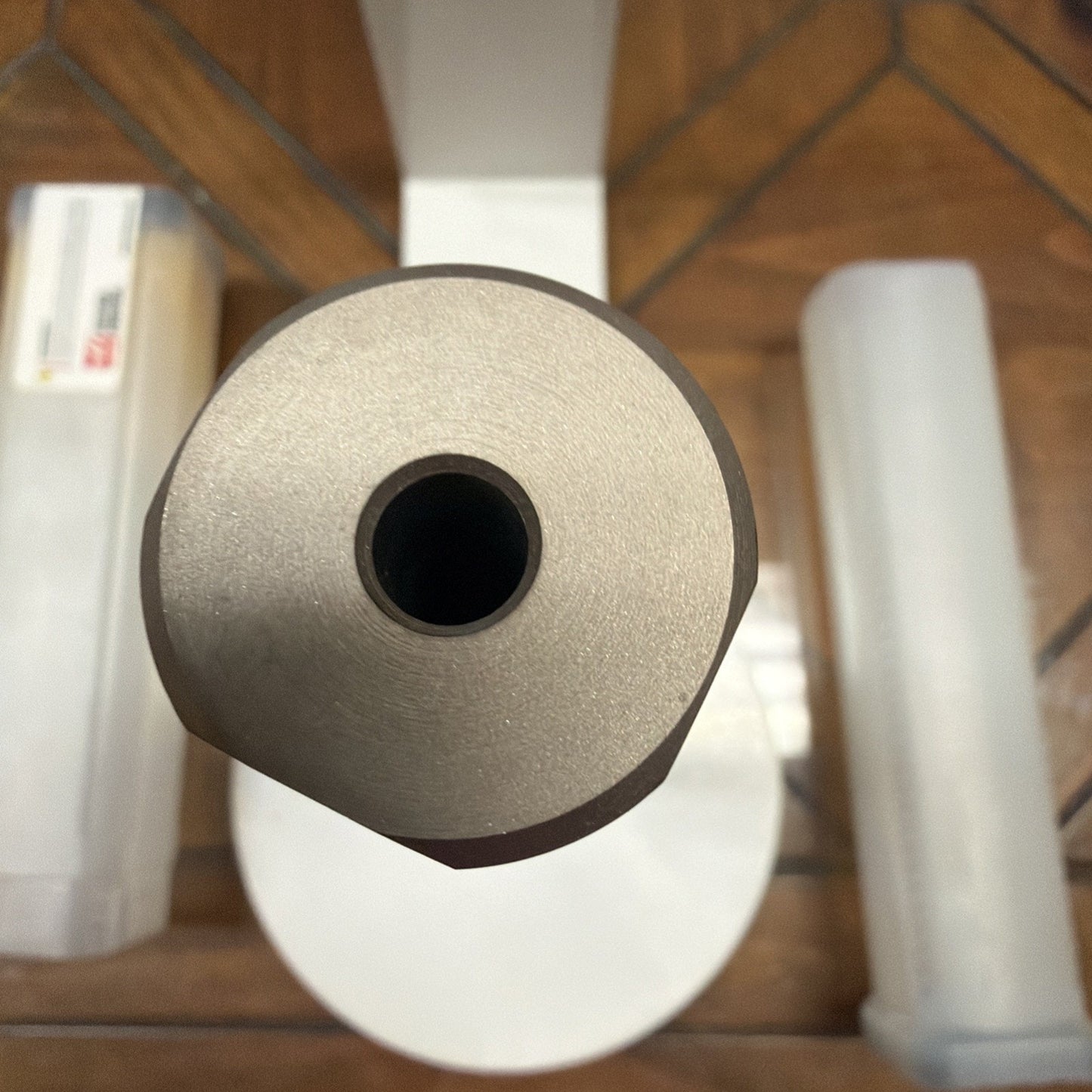 Close-up top view of a metallic cylindrical tool or component with a hollow center, placed on a reflective surface with two rectangular plastic cases nearby on a wooden floor.