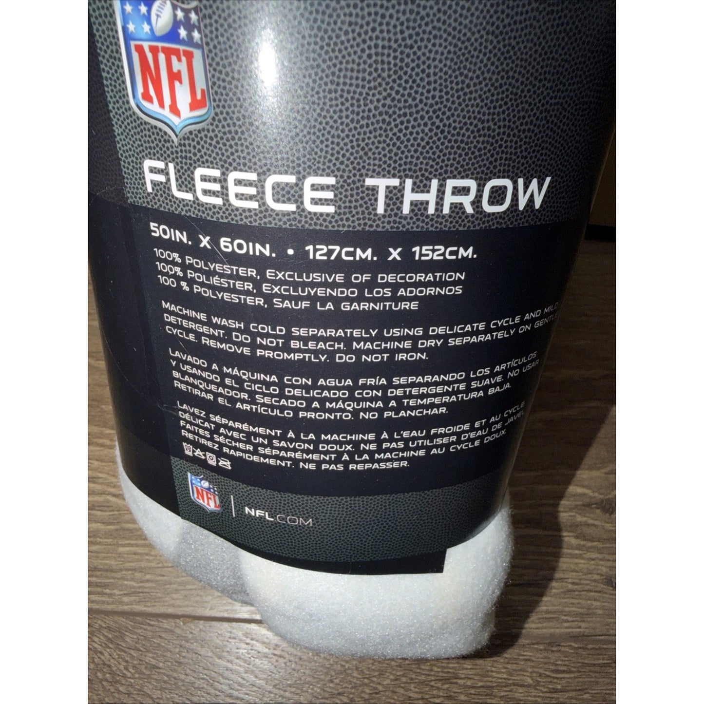 Close-up of a black fleece throw package showing product dimensions, care instructions in multiple languages, NFL logo, and material composition on a wooden surface.
