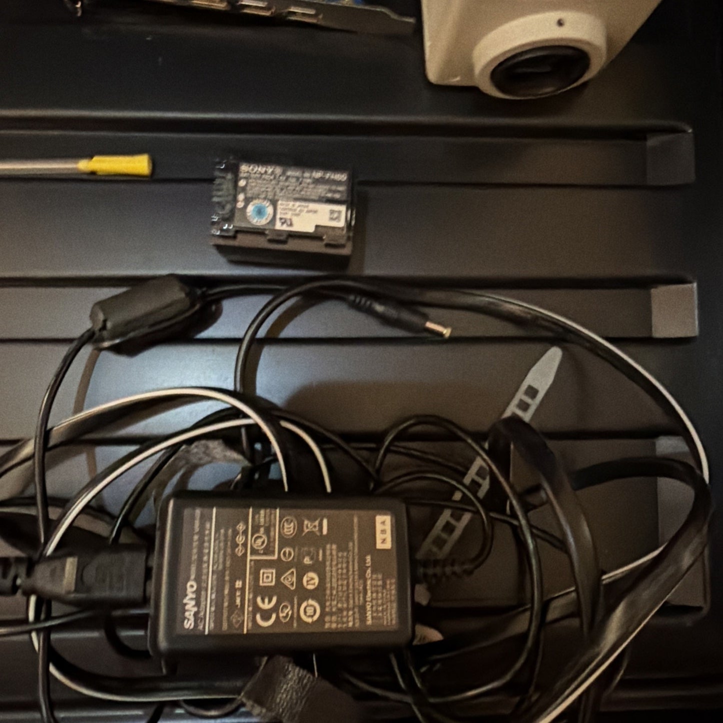 Black Sanyo AC adapter with various certification labels connected to tangled black cables, a small rectangular battery, a yellow-handled tool, and a white dome security camera in the background.