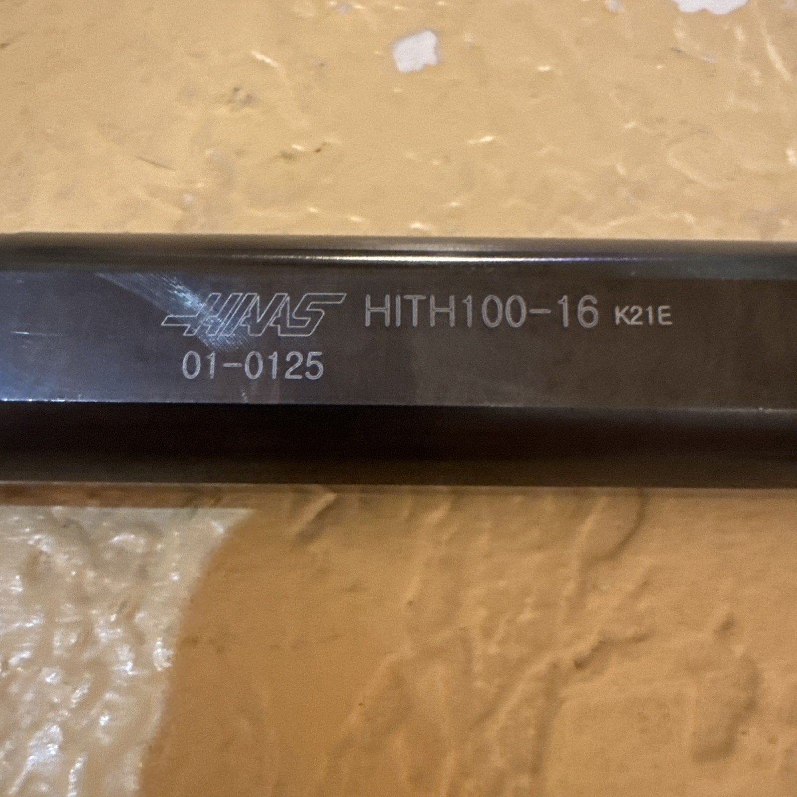 Close-up of a dark metal tool bar displaying engraved text including a brand logo, model number HITH100-16, and additional alphanumeric codes, set against a textured beige background.