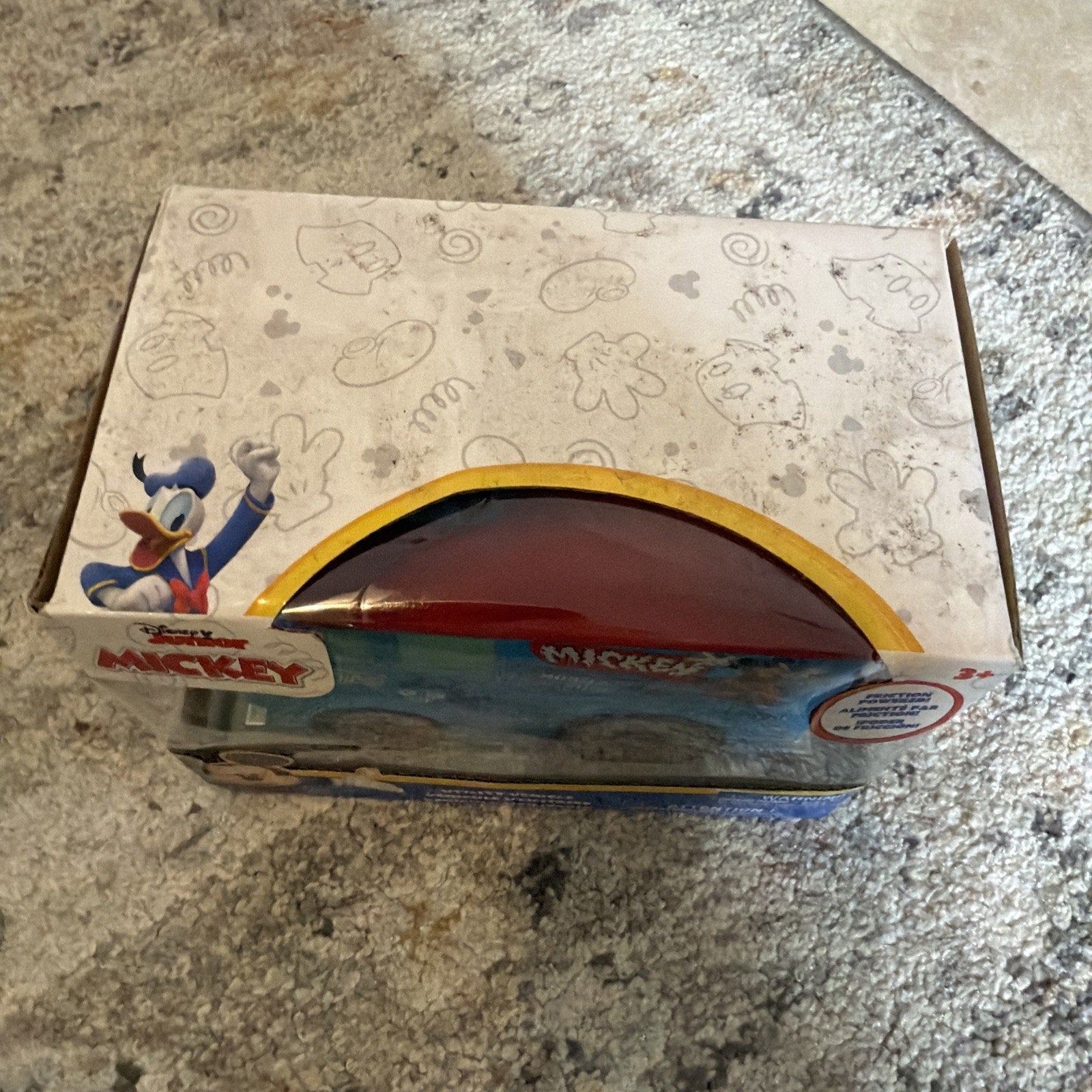 Partially opened rectangular box featuring Disney characters Donald Duck waving on one side and Mickey Mouse-themed illustrations scattered on the white top, resting on a carpeted surface.