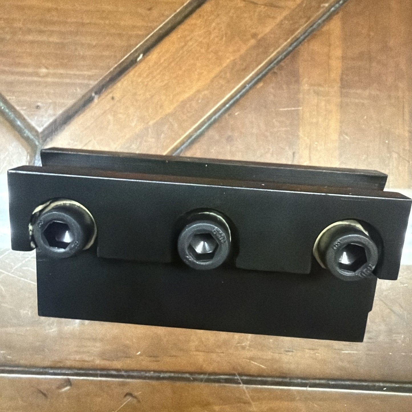 Black metal mechanical component with three hex socket screws aligned horizontally, placed on a wooden surface with visible grain patterns.