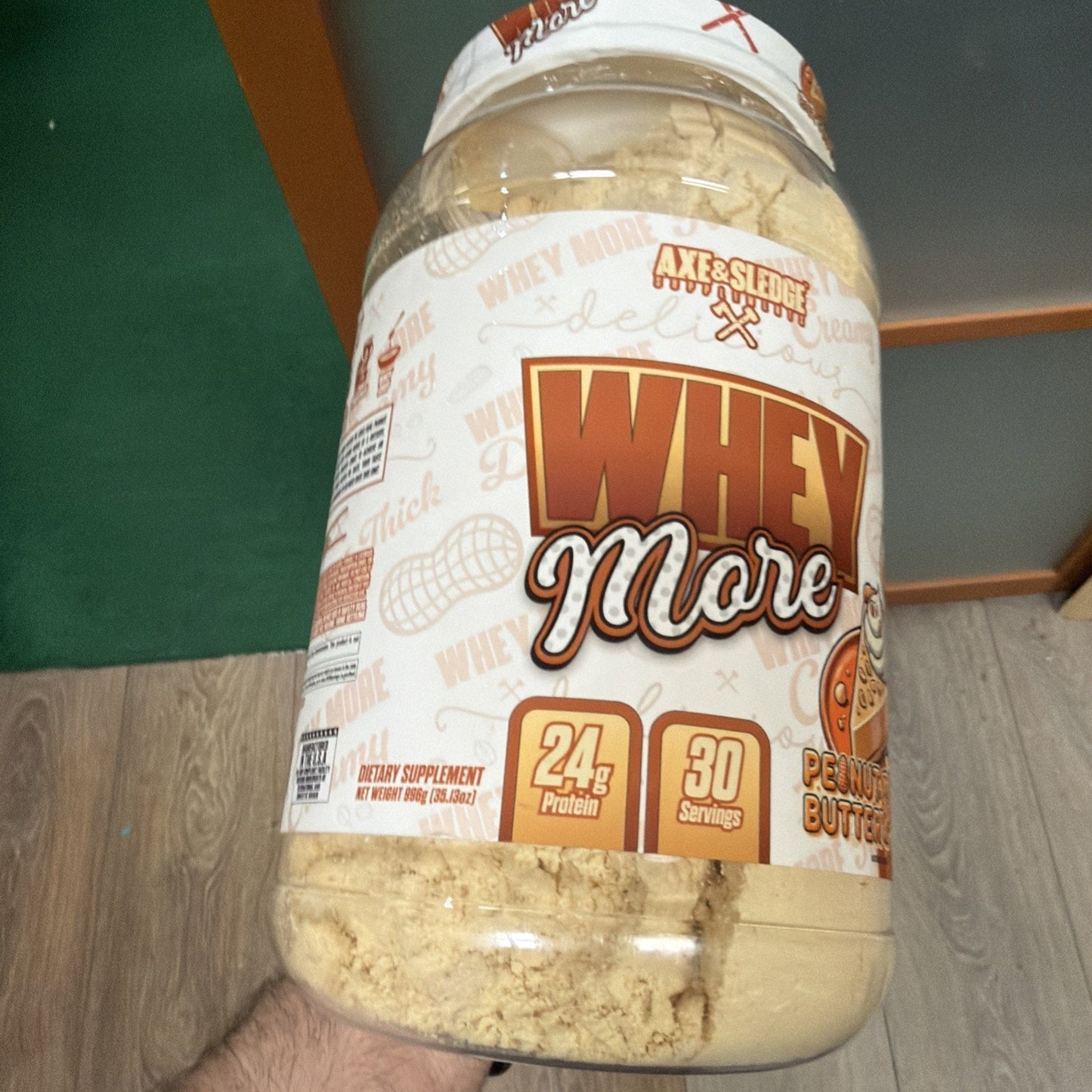 Large clear plastic container of peanut butter-flavored whey protein powder with a label highlighting 24 grams of protein and 30 servings per container, held by a hand over wooden flooring.