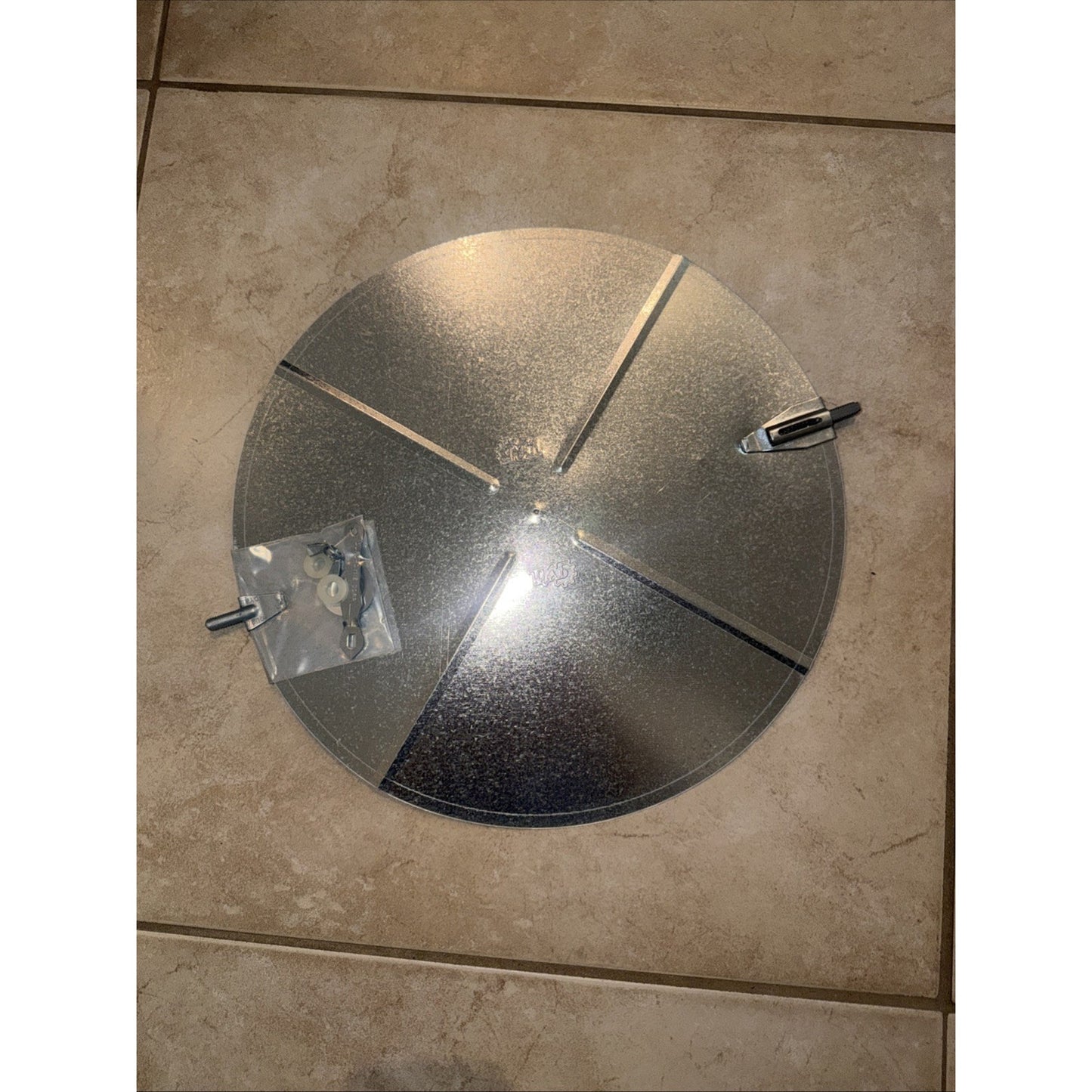 Round galvanized metal vent cover with adjustable slats and two mounting clips, placed on a tiled floor next to a small plastic bag containing screws and hardware.