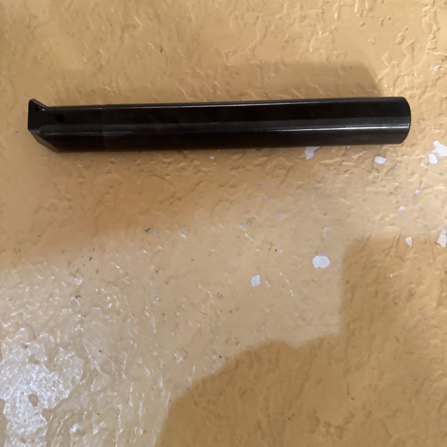 Long black cylindrical metal tool with a flat cutting edge at one end, resting horizontally on a textured beige surface with scattered white spots.