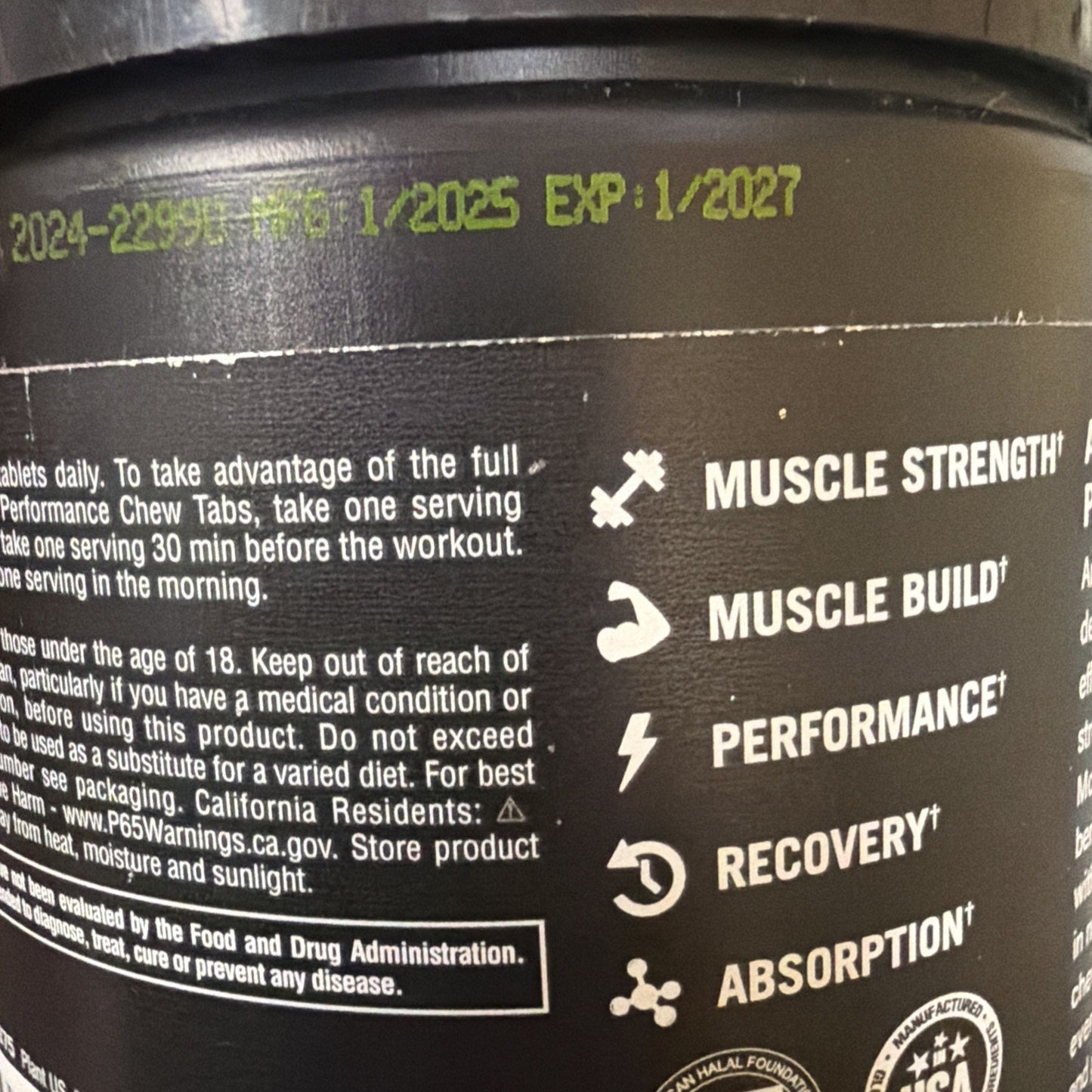 Close-up of a black plastic supplement container showing yellow printed manufacturing date 1/2025, expiration date 1/2027, usage directions, and icons representing muscle strength, muscle building, performance, recovery, and absorption benefits.