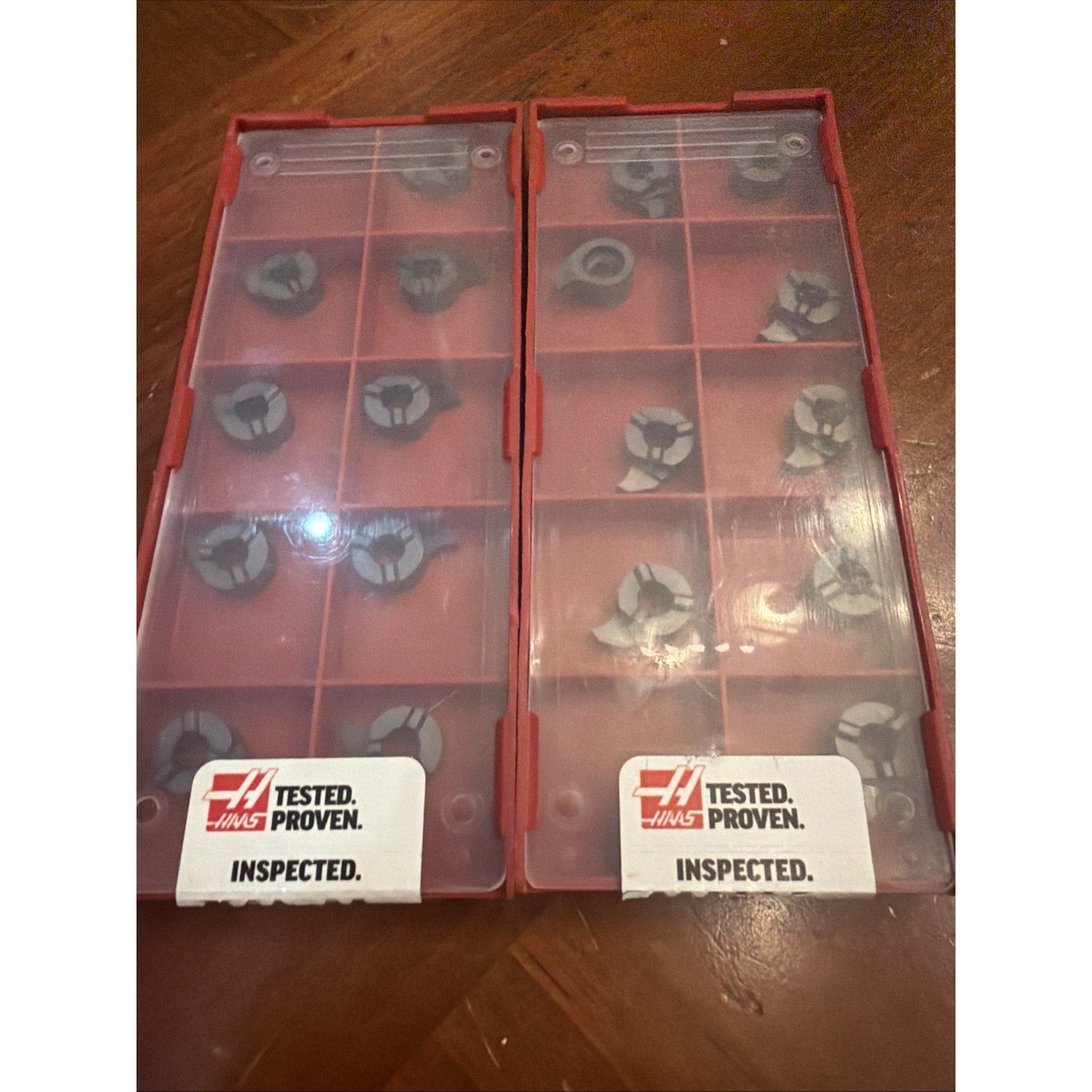 Two red plastic storage cases with translucent lids, each containing multiple small three-pronged metal inserts, placed side by side on a wooden surface with "Haas Tested Proven Inspected" labels visible on both cases.