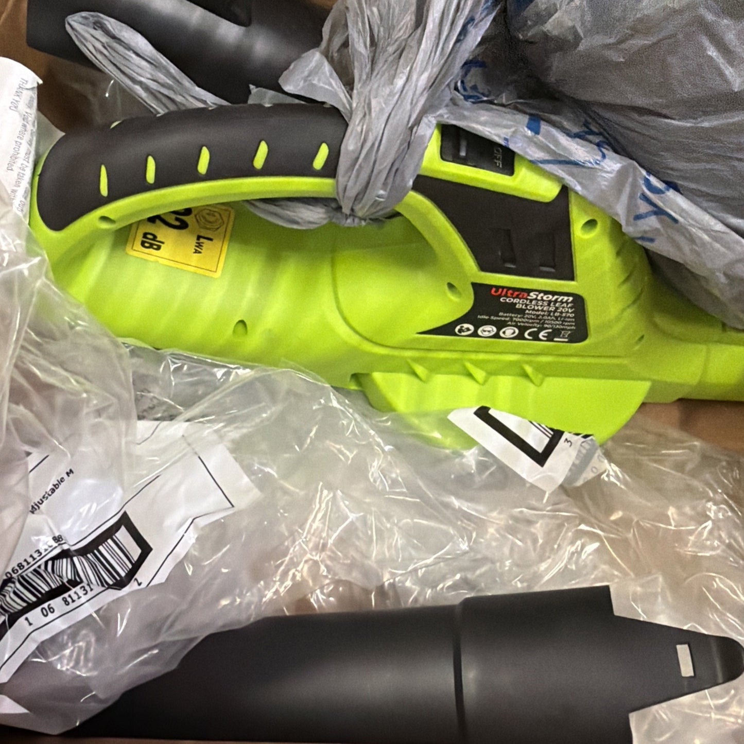 Close-up of a bright green handheld cordless leaf blower with black ergonomic grip and attached label, partially wrapped in plastic and surrounded by packaging materials.