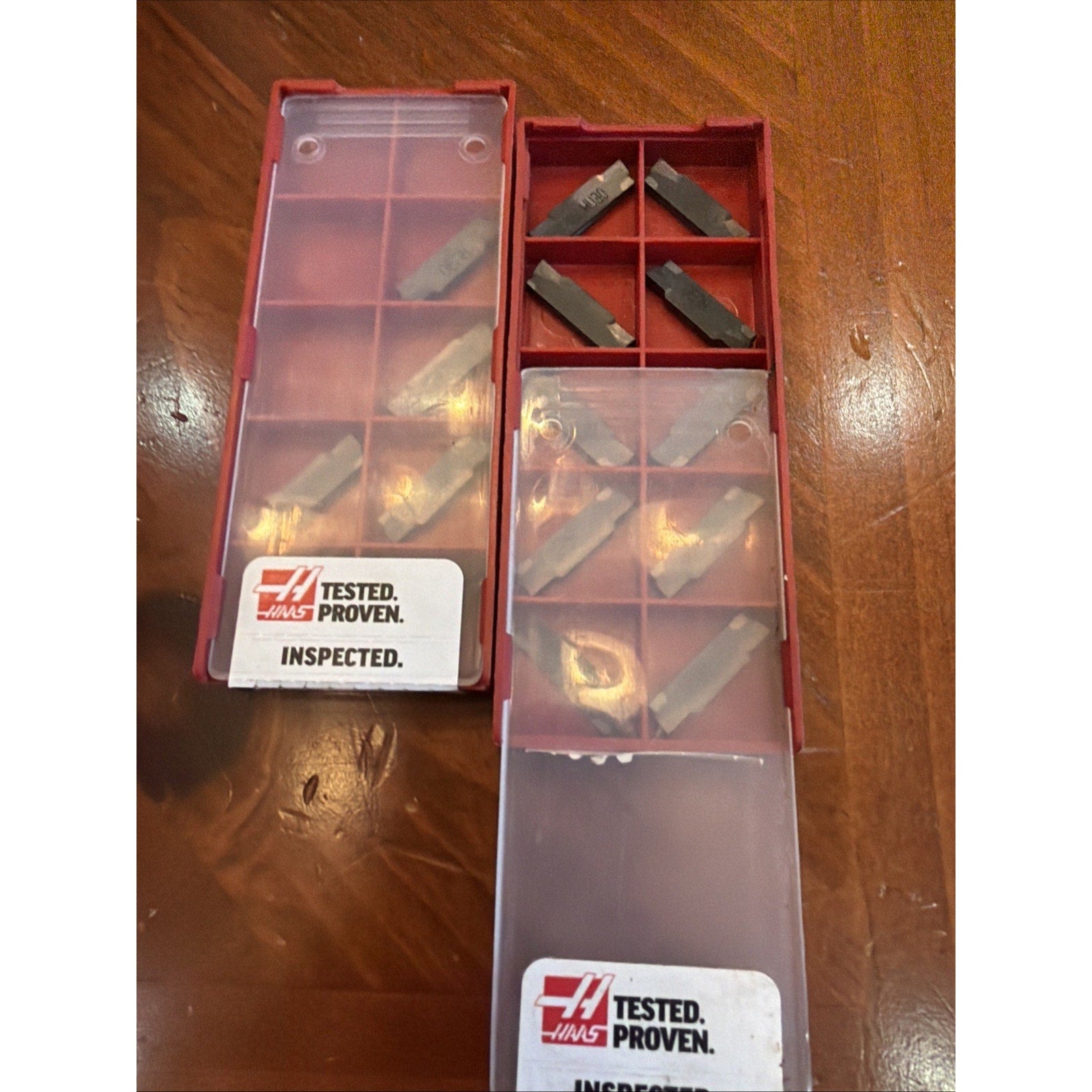 Two red plastic storage boxes on a wooden surface, each containing several dark metal cutting inserts in individual compartments, with translucent sliding lids and Haas Tested Proven Inspected labels.