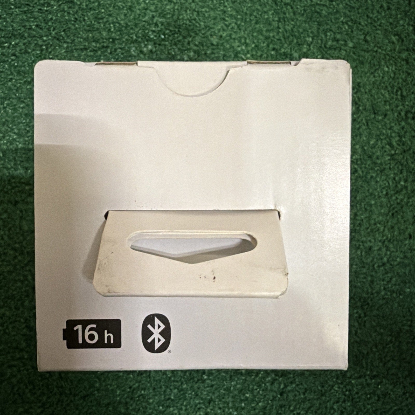 White rectangular cardboard box with a cutout handle on one side, featuring icons indicating 16-hour battery life and Bluetooth connectivity, placed on a green textured surface.