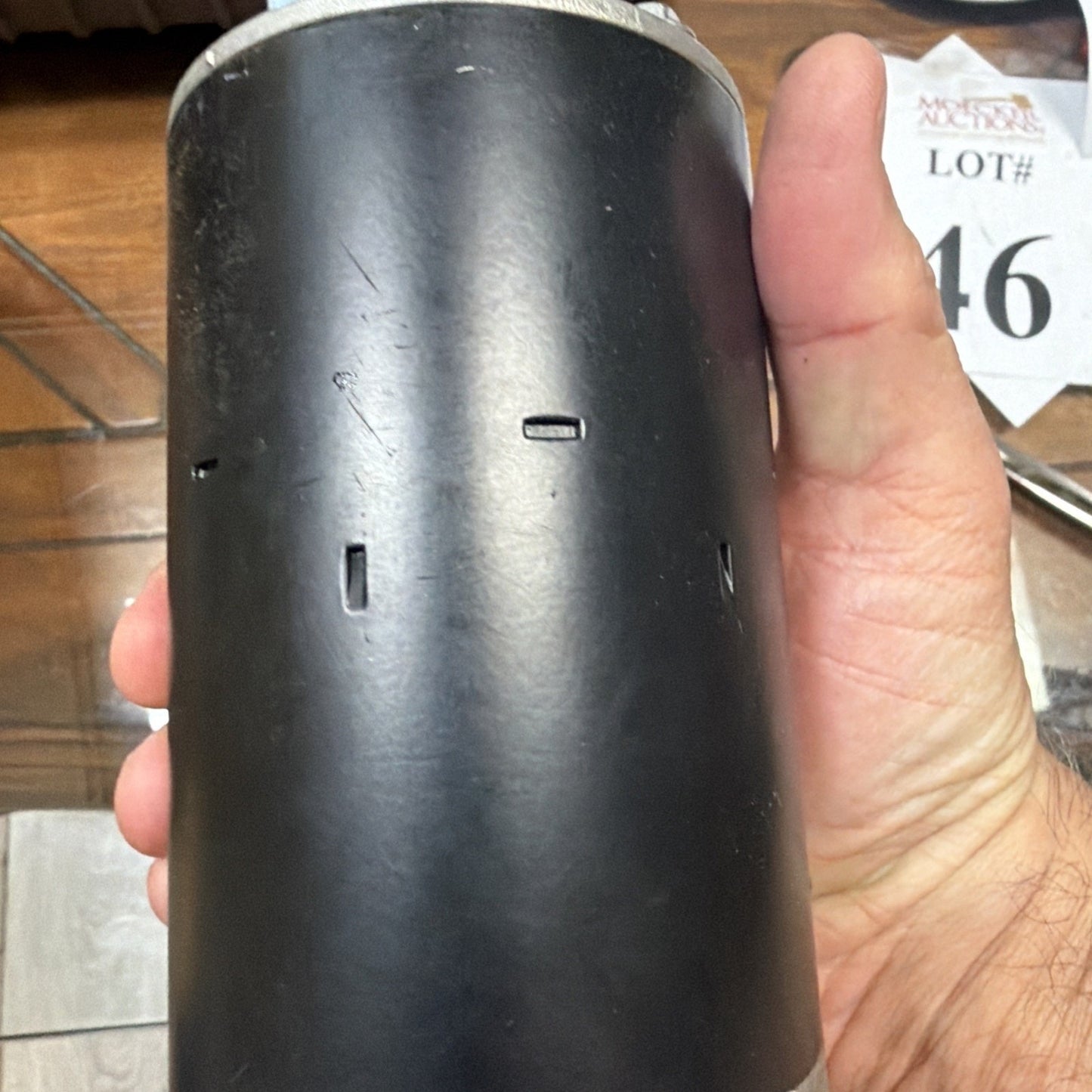 Hand holding a cylindrical black metal object with three rectangular cutouts, resting on a wooden floor with a white auction lot card visible in the background.