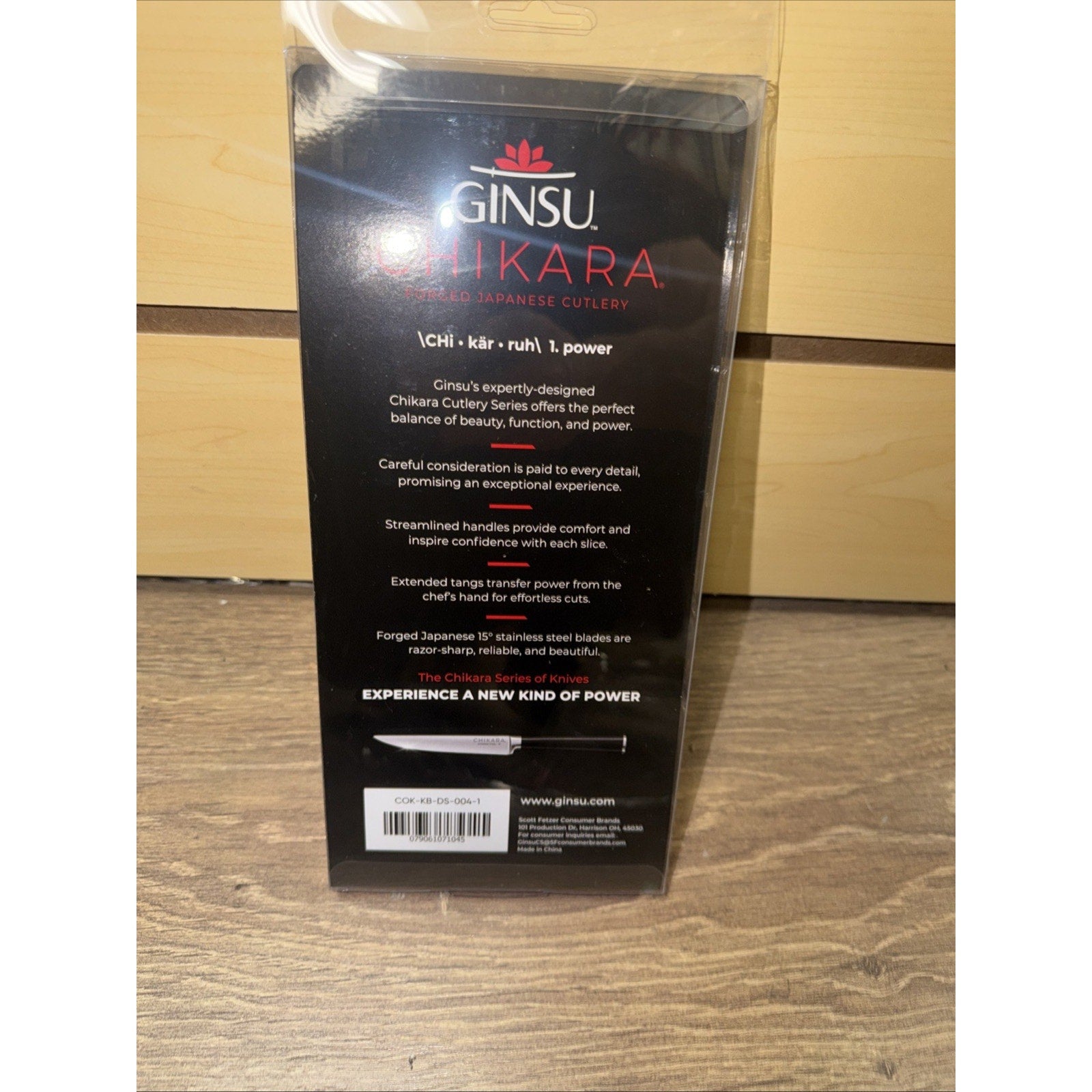 Back view of Ginsu Chikara cutlery packaging showing product description highlighting design, balance, comfort, forged Japanese steel blades, and the brand website on a wooden surface background.