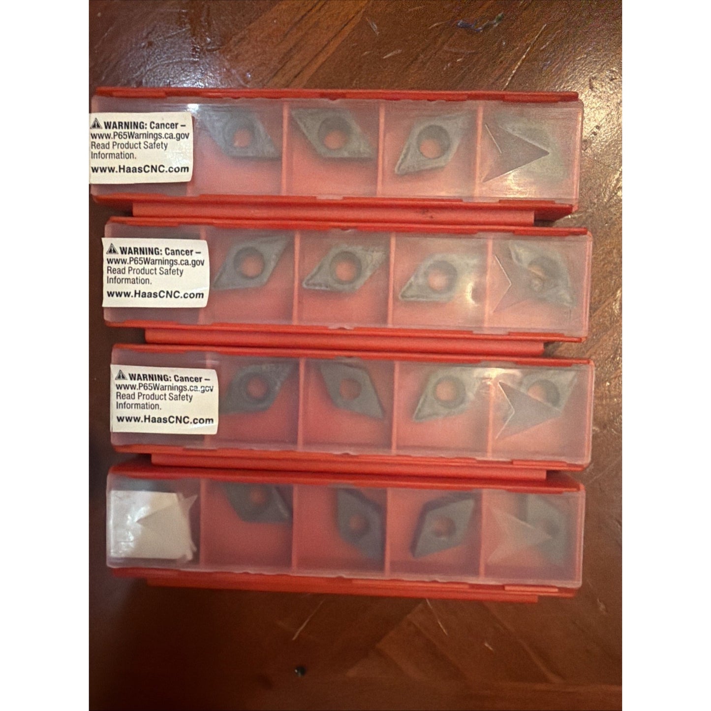 Four stacked red plastic containers each divided into compartments holding various small metal cutting inserts, with warning labels visible on some containers, resting on a wooden surface.