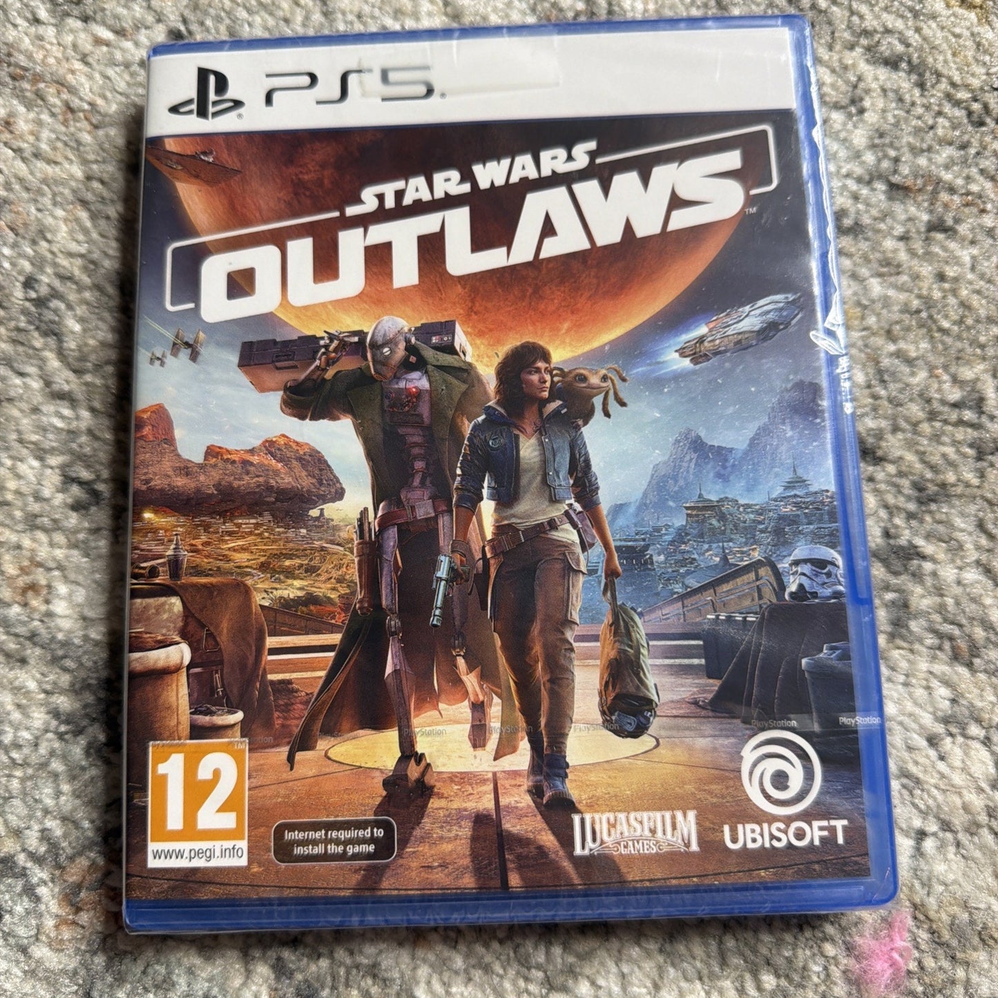 PlayStation 5 game case for Star Wars Outlaws featuring characters and spacecraft against a fiery planet backdrop, with PEGI 12 rating and logos for Lucasfilm Games and Ubisoft.