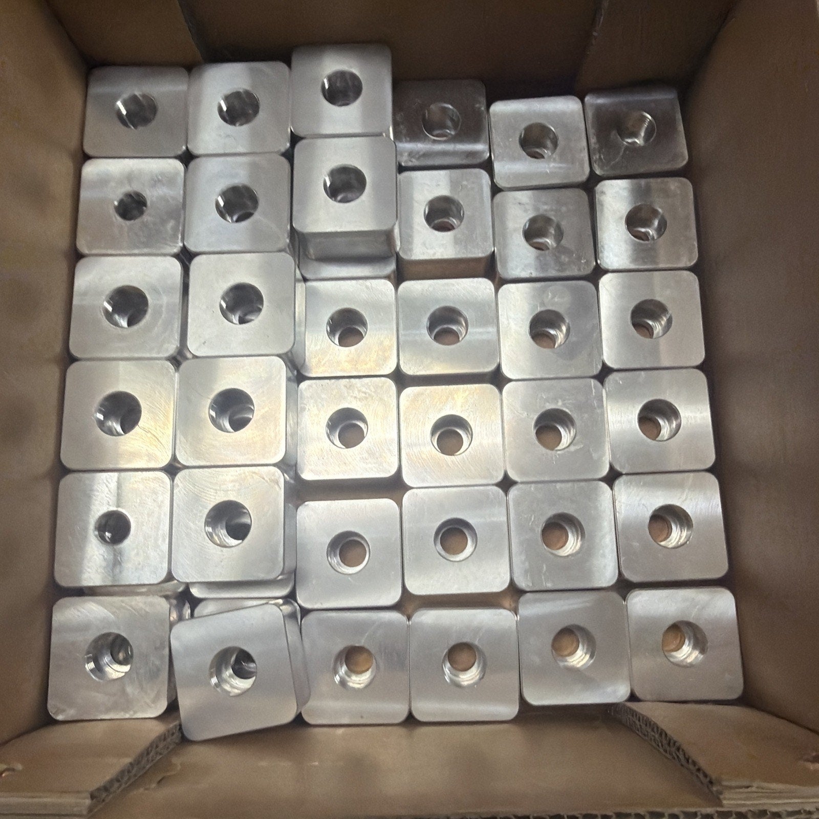 Cardboard box filled with multiple stacked square metal pieces each featuring a central circular hole, arranged in neat rows.