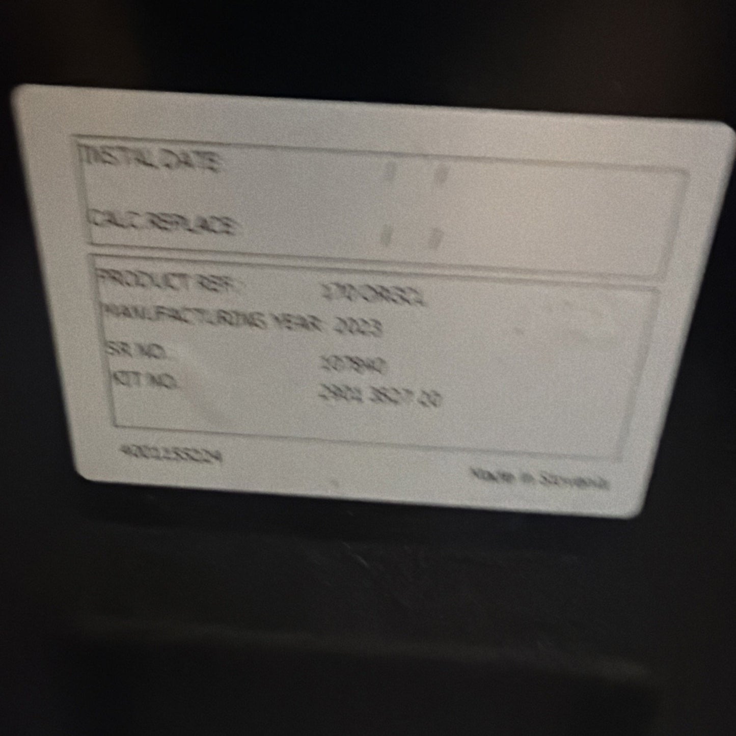 Blurred white label on a dark surface showing faint printed text including product item details, manufacturing year 2023, serial number, and "Made in Standard.