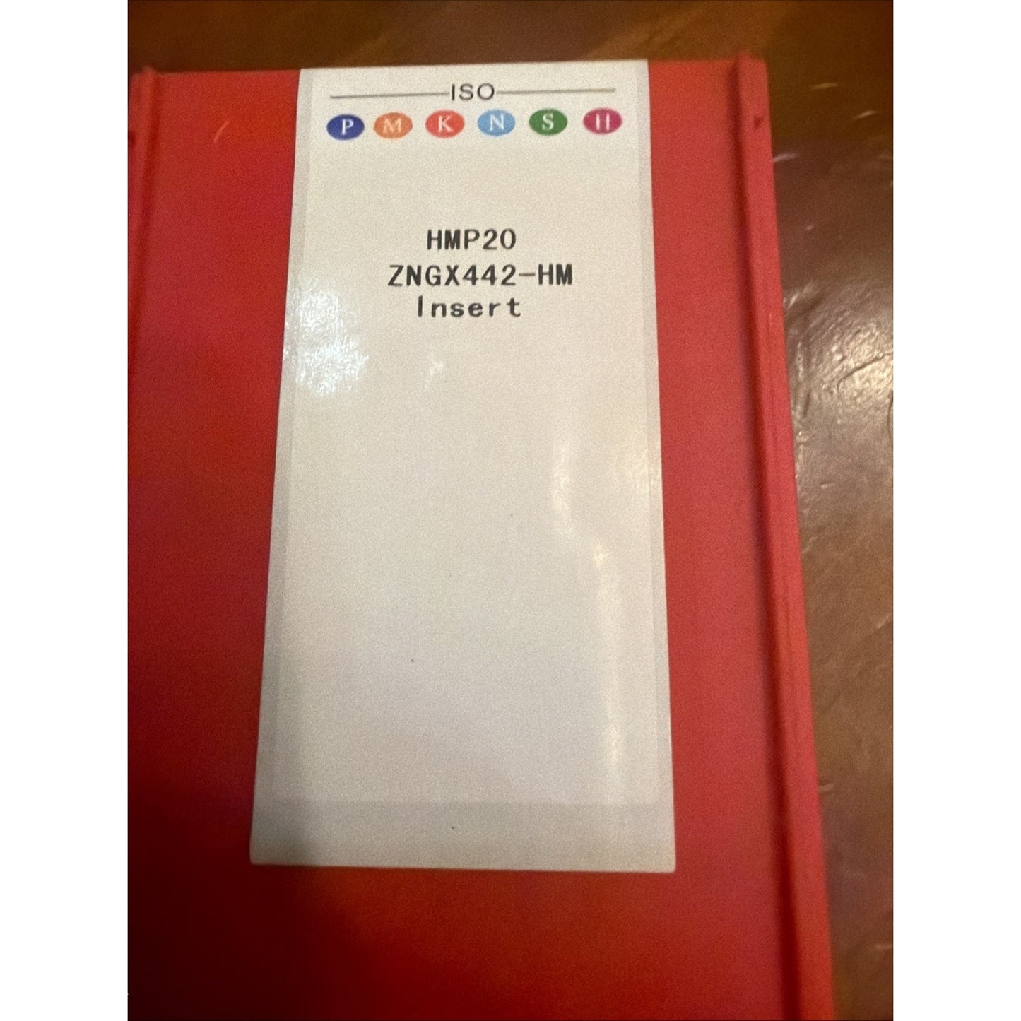 Red rectangular plastic container with a white label showing text "ISO," colored circles, and product code "HMP20 ZNGX442-HM Insert," placed on a wooden surface.