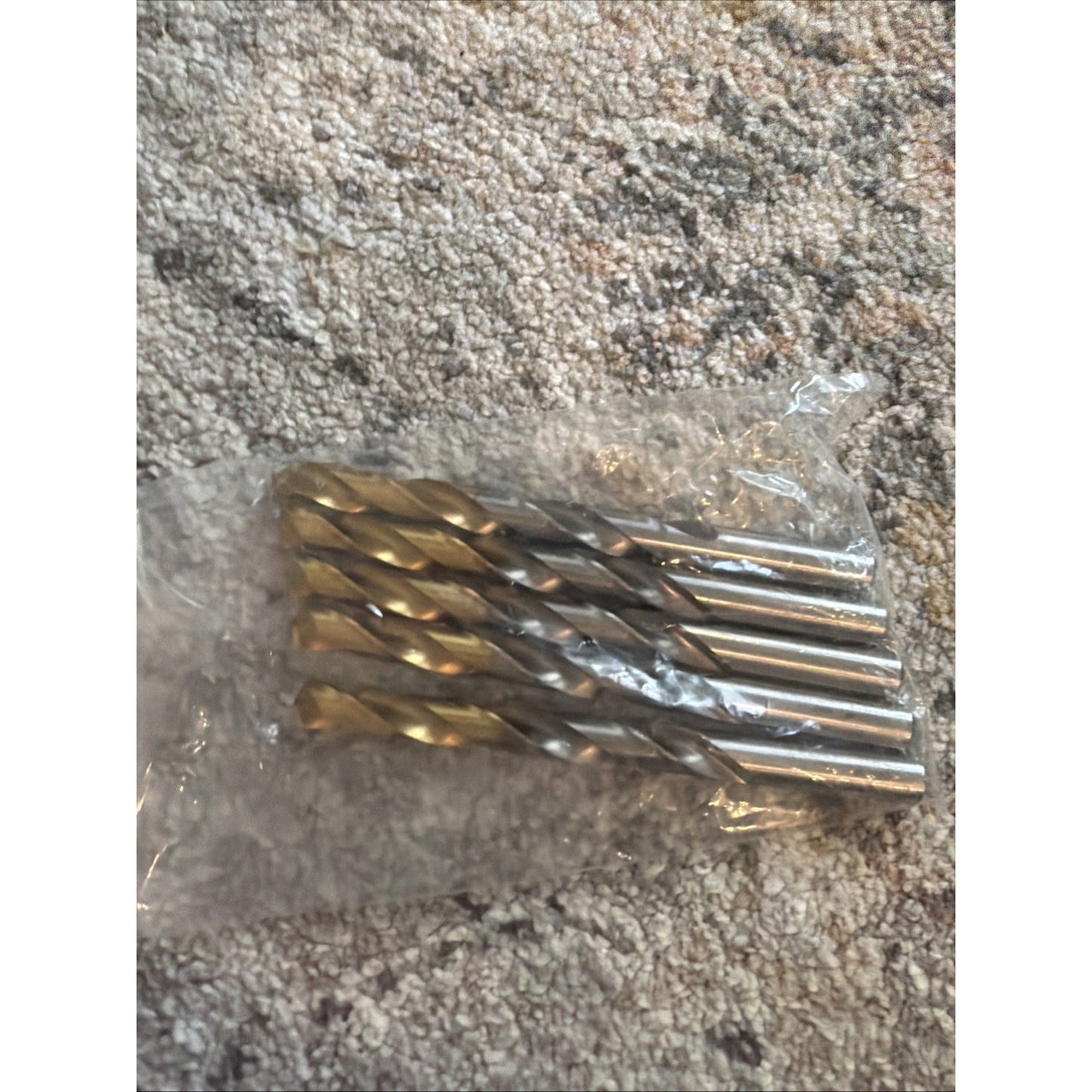 Set of four metal twist drill bits with spiral flutes, packaged together in a clear plastic bag, placed on a textured carpeted surface.