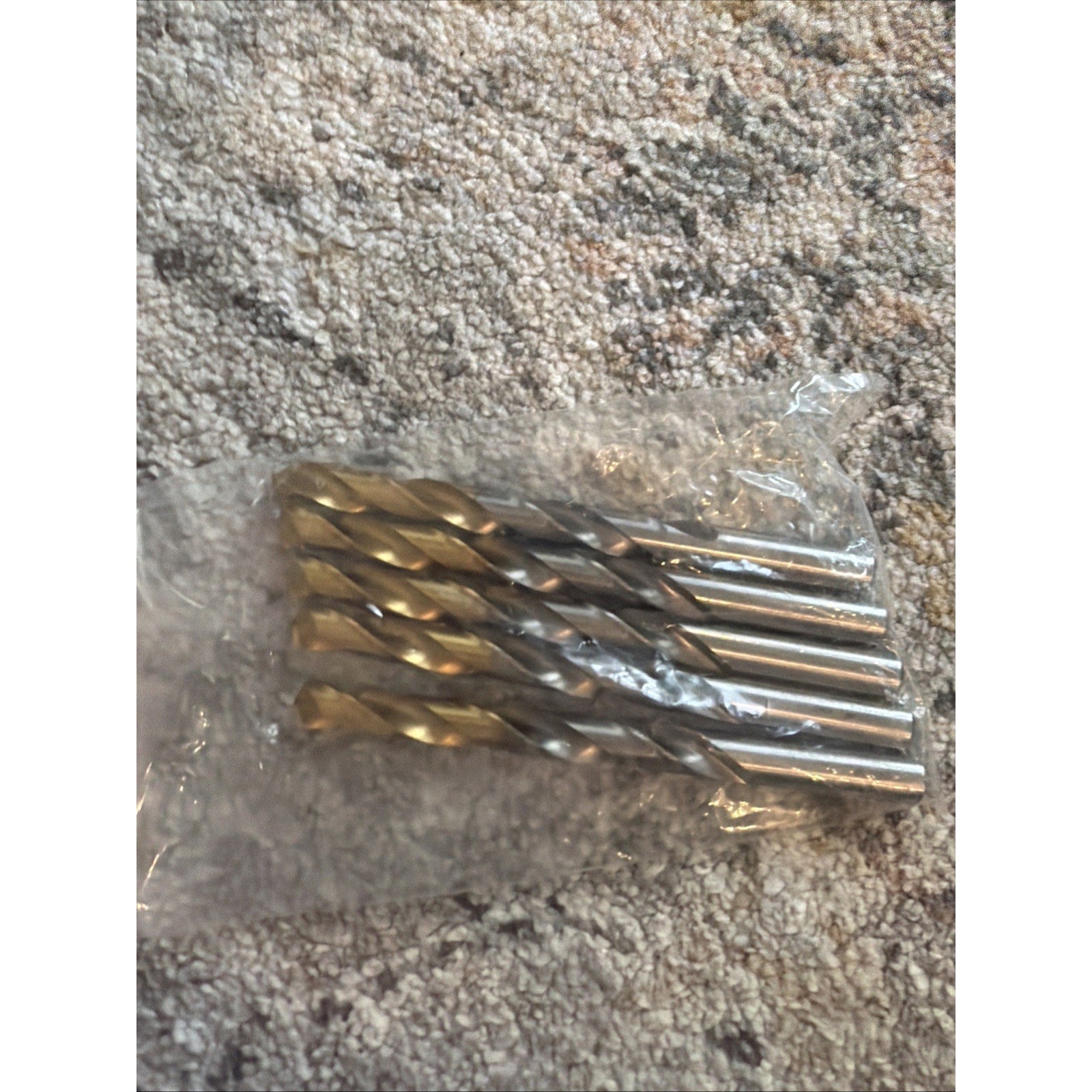 Set of four metal twist drill bits with spiral flutes, packaged together in a clear plastic bag, placed on a textured carpeted surface.