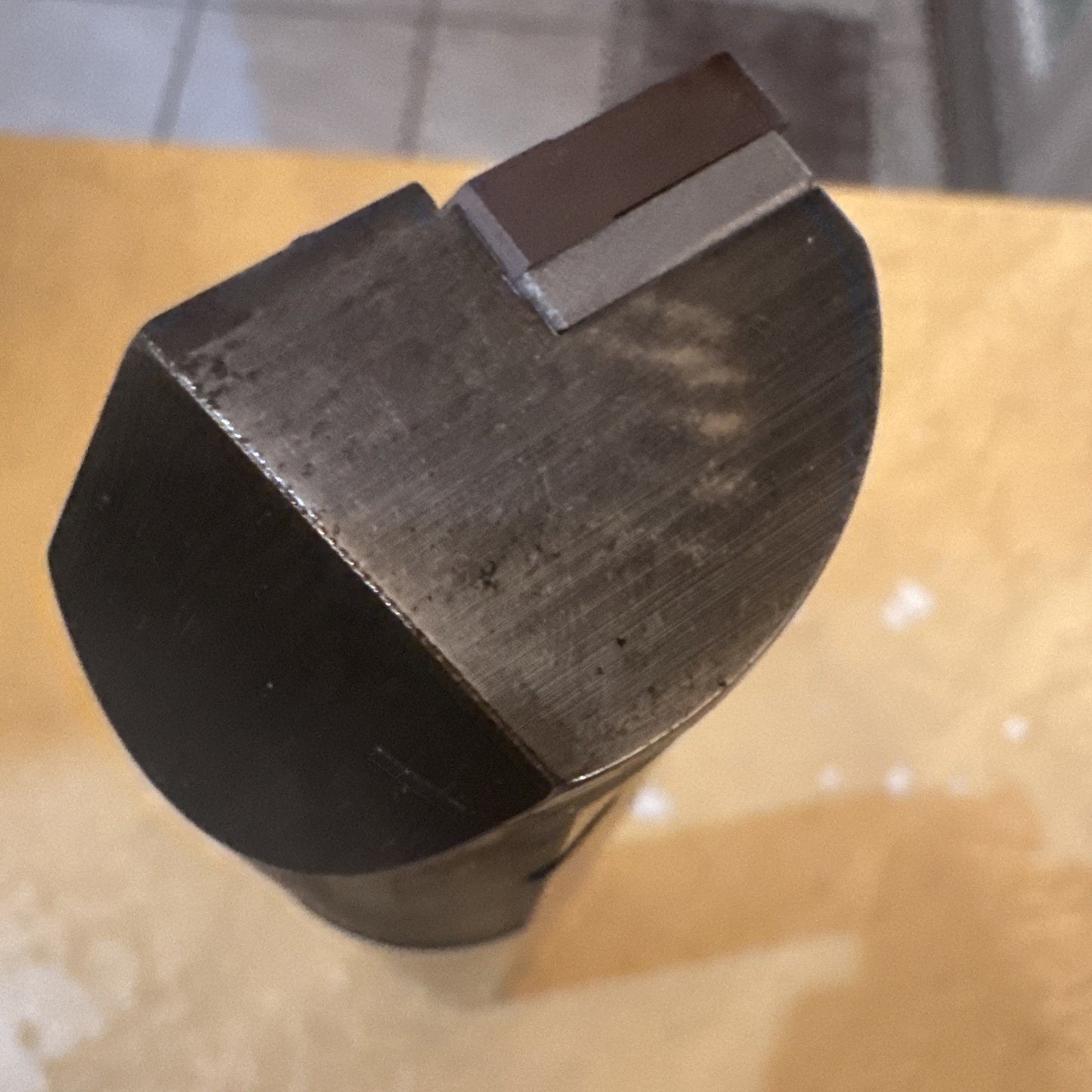 Close-up of a dark metal cutting tool tip with a flat semi-circular face and a small rectangular raised section near the edge, placed on a yellowish surface.