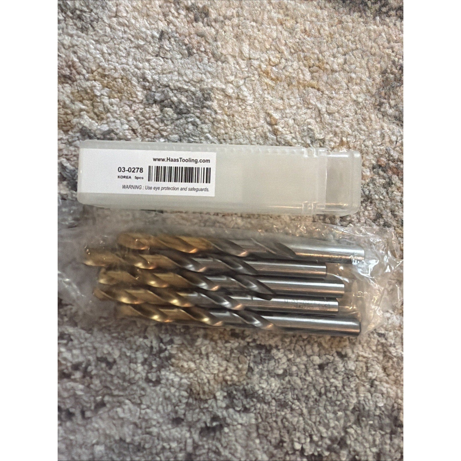 Set of five metal twist drill bits with gold-colored tips and silver shanks, packed together in clear plastic, placed next to a white plastic box with a barcode label on a textured carpet surface.