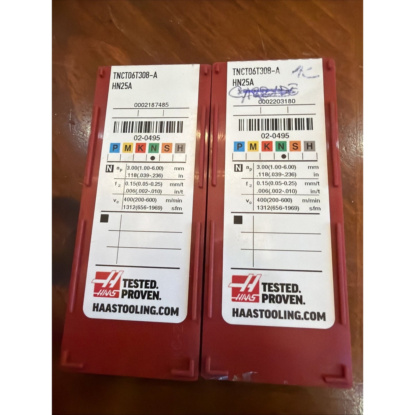 Two red plastic cases with white labels showing technical machining parameters, colored ISO classification codes, barcodes, and Haas branding, placed side by side on a wooden surface.