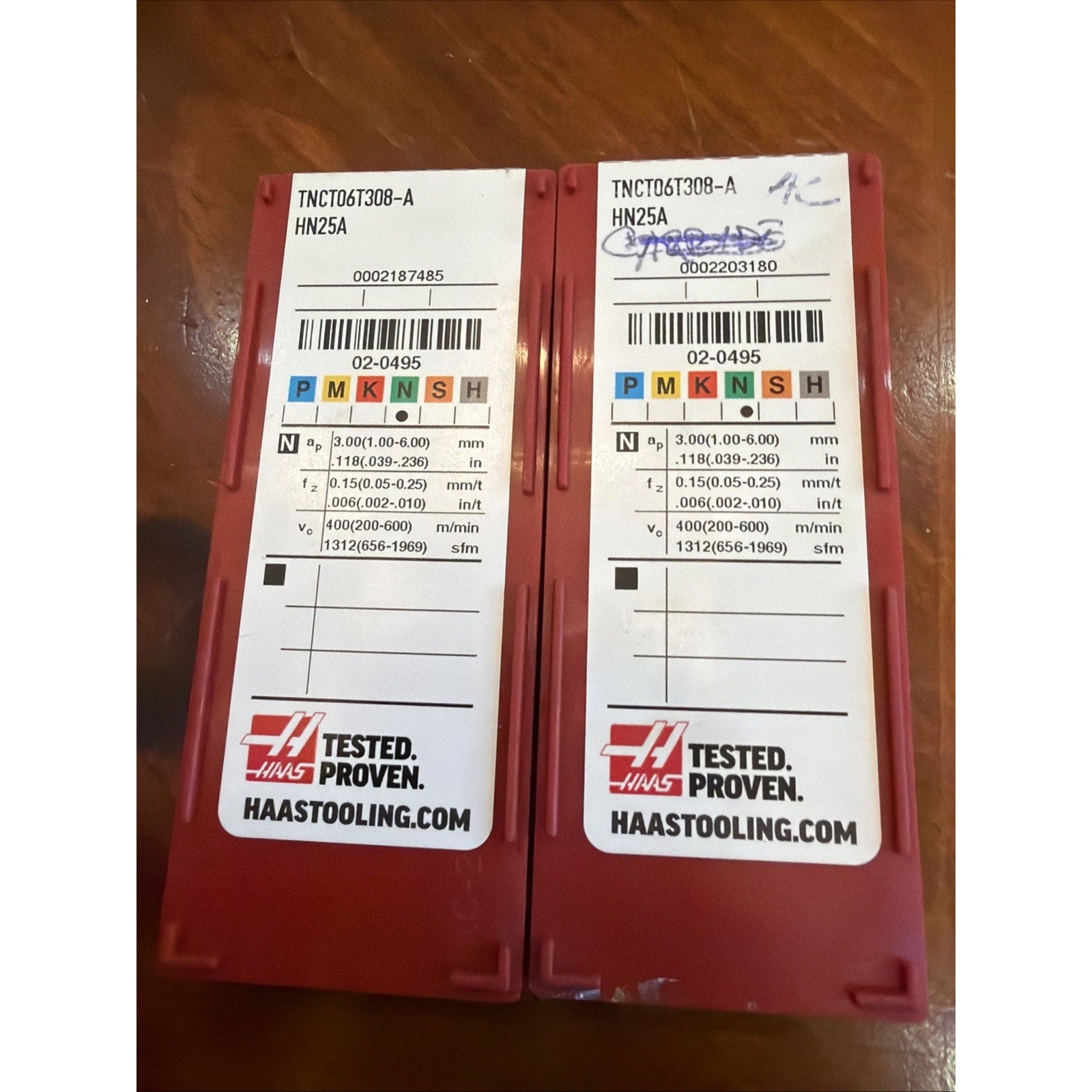 Two red plastic cases with white labels showing technical machining parameters, colored ISO classification codes, barcodes, and Haas branding, placed side by side on a wooden surface.