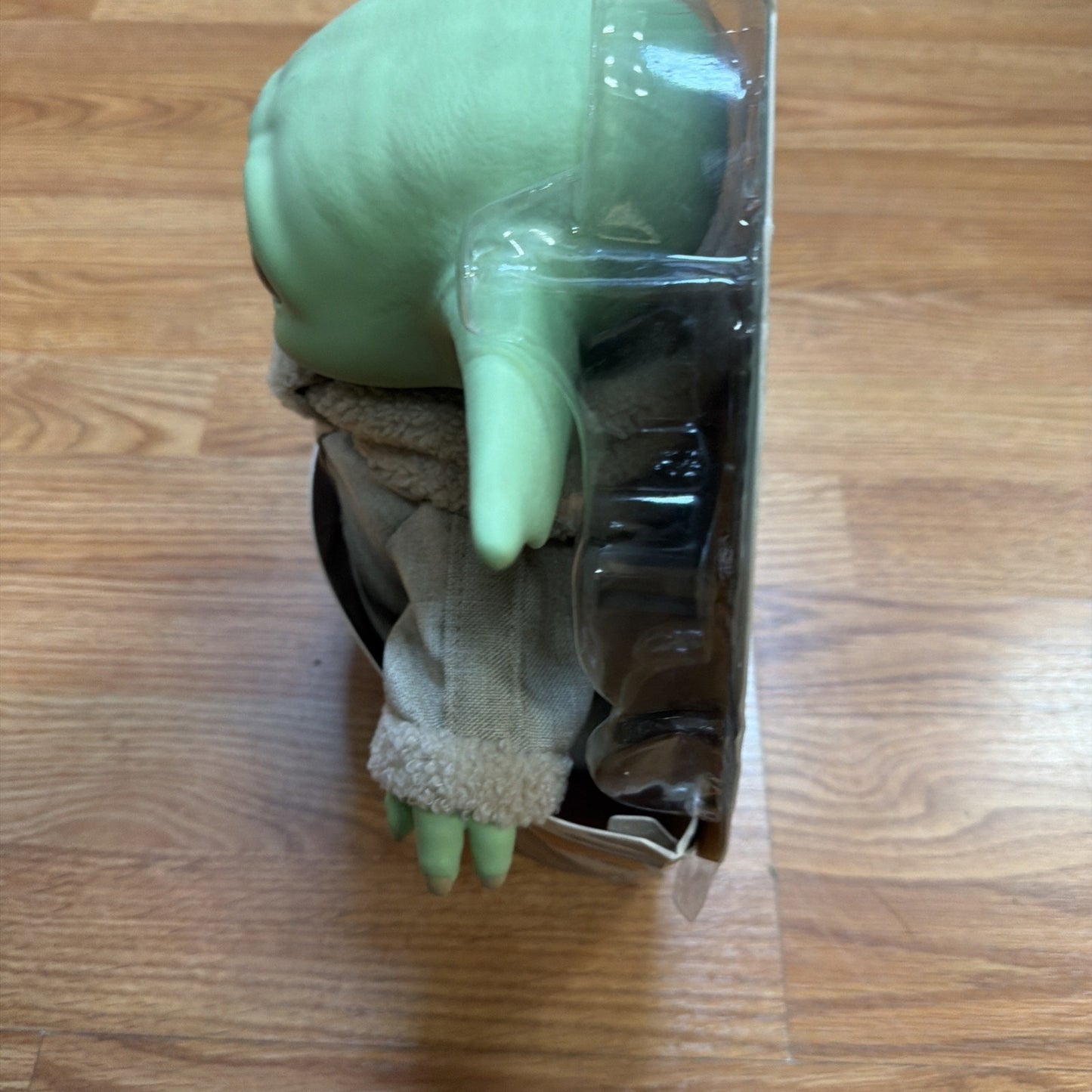 Side view of a green, alien-like figure with large ears wearing a beige fleece jacket inside clear plastic packaging on a wooden surface.