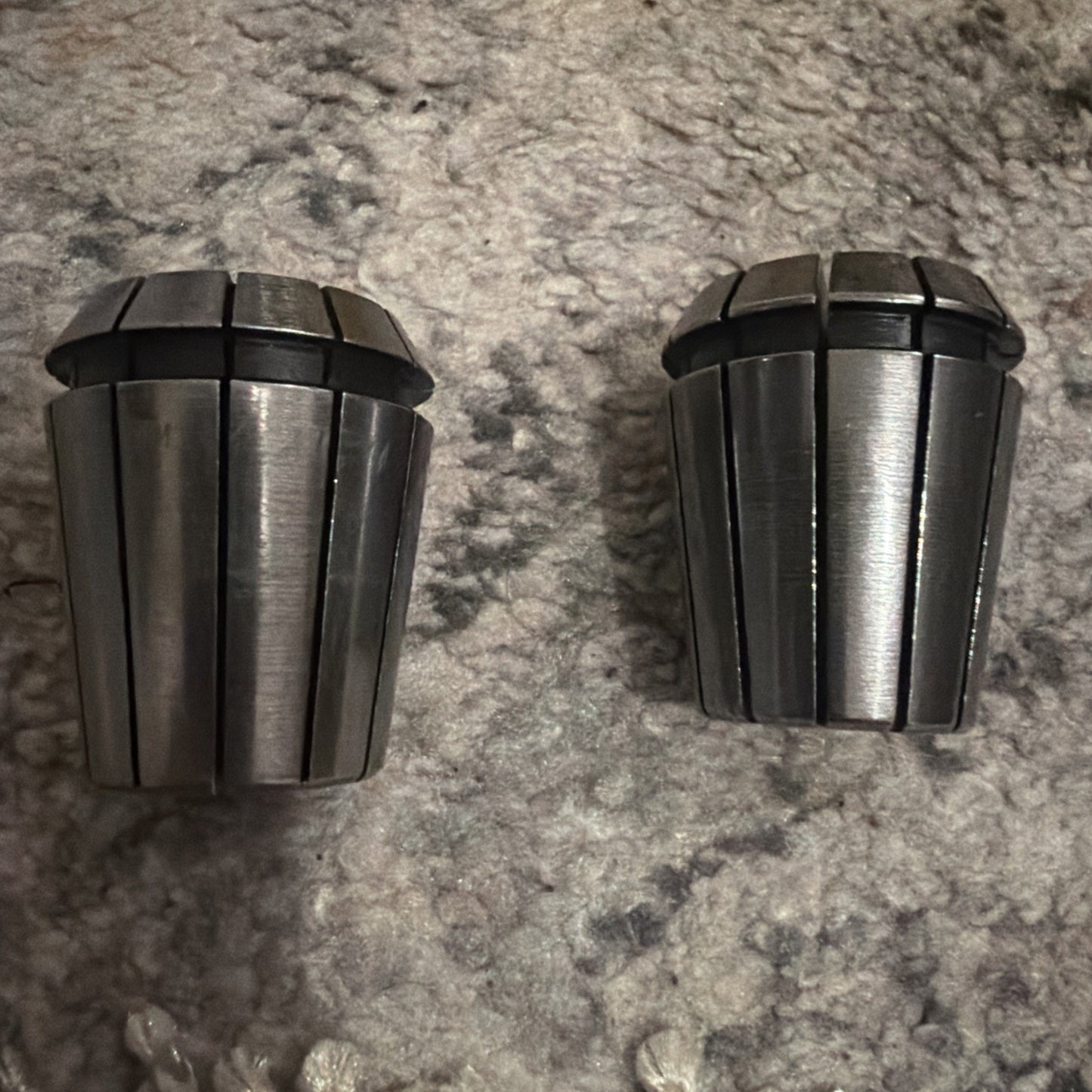 Two shiny metal collets with multiple vertical slits and black rubber rings near the top, placed on a textured gray and beige carpet surface.