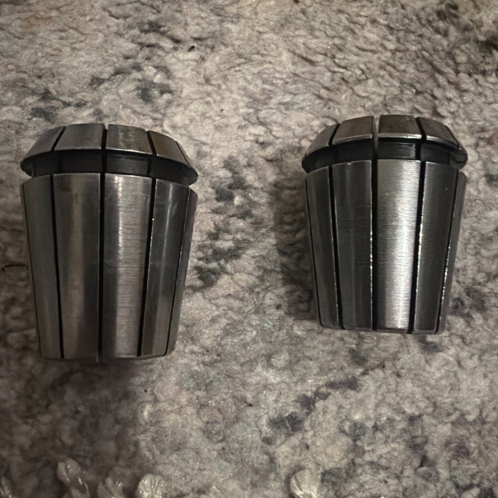 Two shiny metal collets with multiple vertical slits and black rubber rings near the top, placed on a textured gray and beige carpet surface.