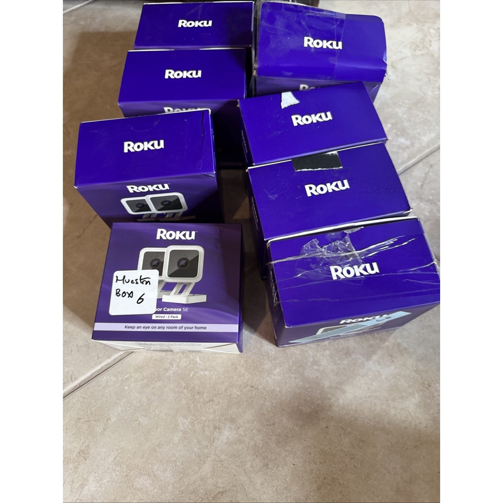 Stack of Roku Indoor Camera SE Wired 2 Pack boxes in purple packaging arranged on tile floor for home surveillance use